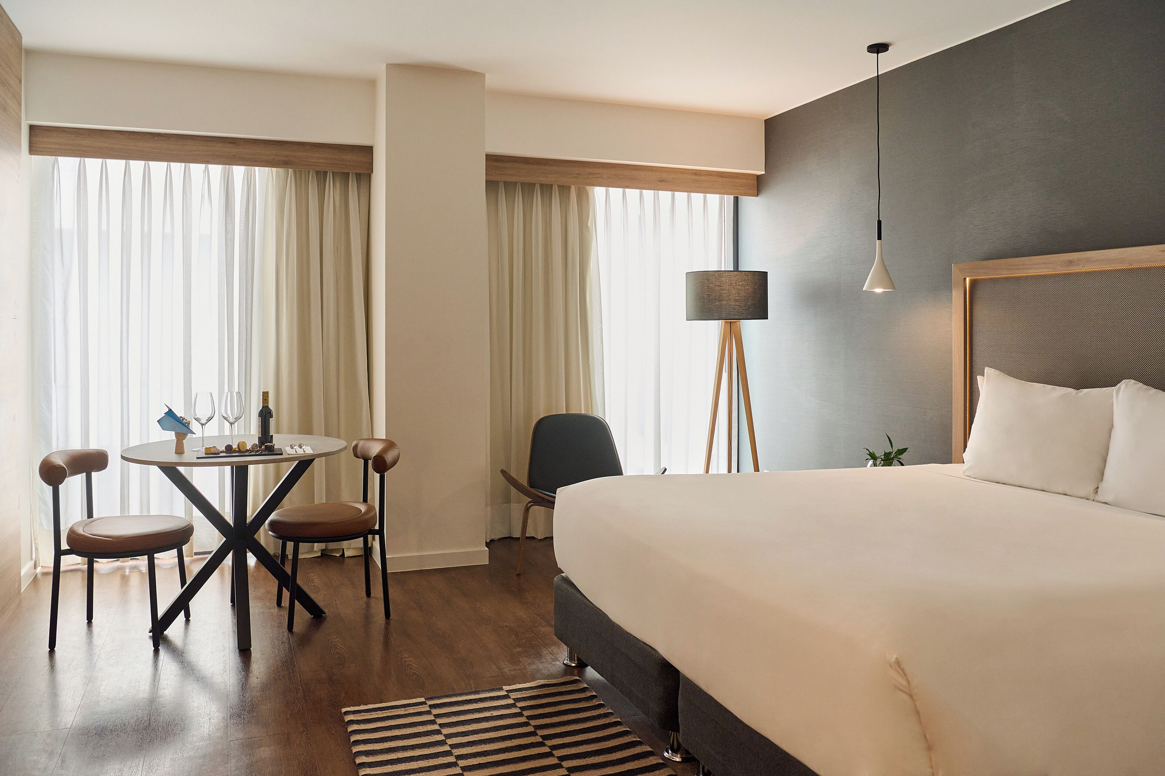 Een hotelkamer met een groot bed, een eettafel met stoelen, een vloerlamp en uitzicht op het landschap door dunne gordijnen.