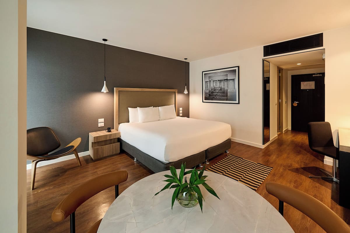 Quarto de hotel moderno com cama grande, mesas de cabeceira, uma mesa de jantar e uma planta em vaso.