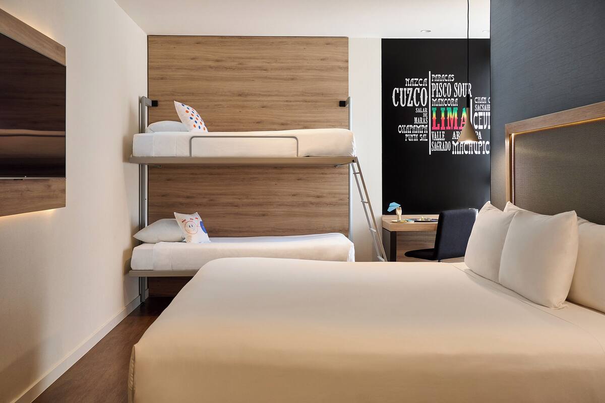 Quarto de hotel com beliche, escrivaninha e uma cadeira, além de uma obra de arte na parede.