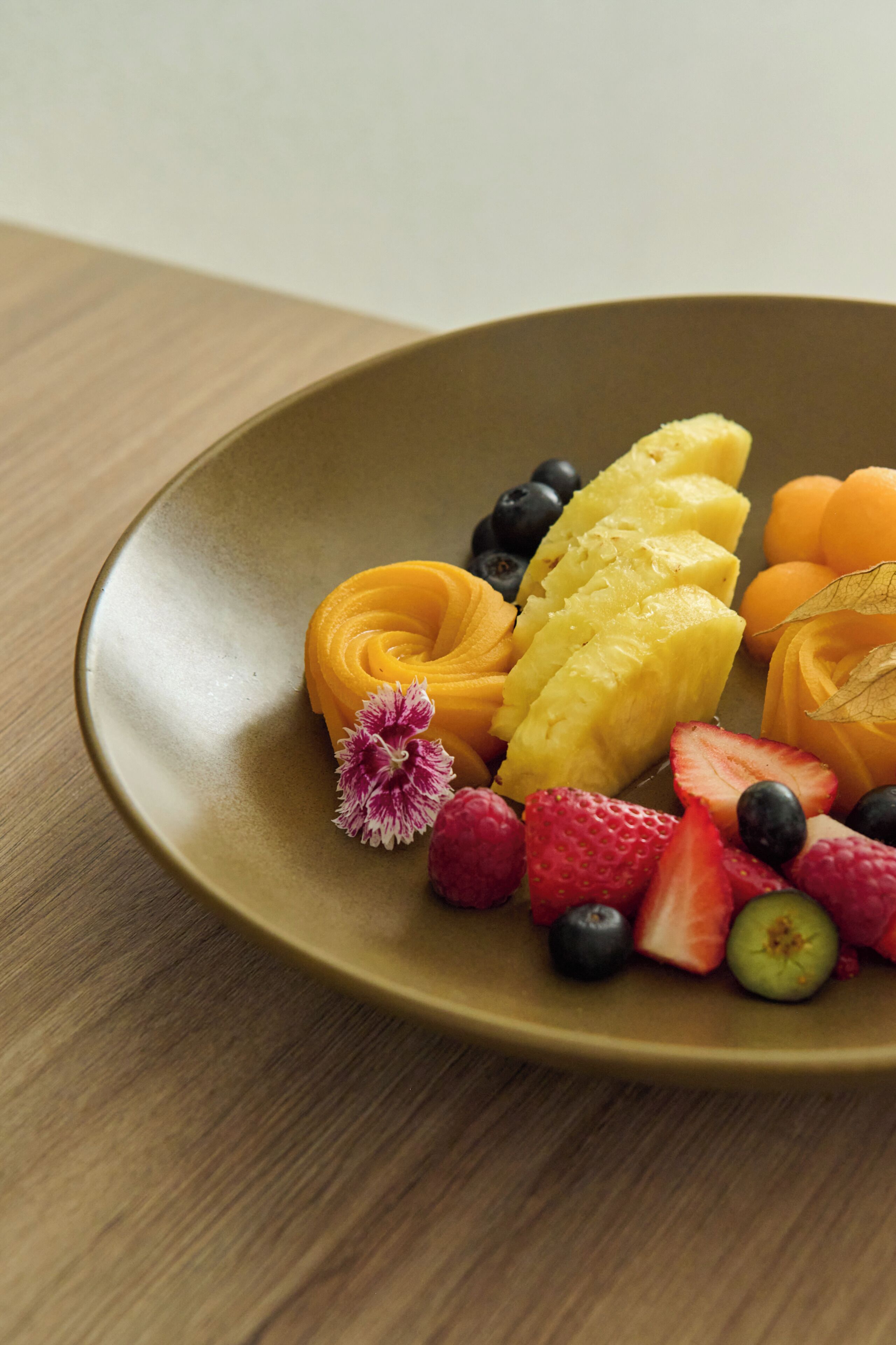 A round, brown plate with various fruits including pineapple, mango, strawberries, blueberries, and raspberries.