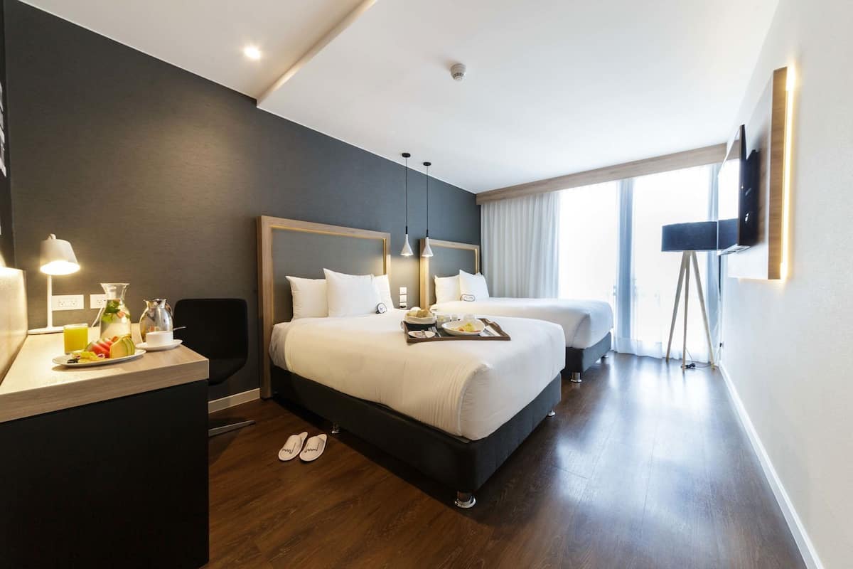 Quarto de hotel com duas camas, uma escrivaninha com luminária, televisão e uma janela grande.