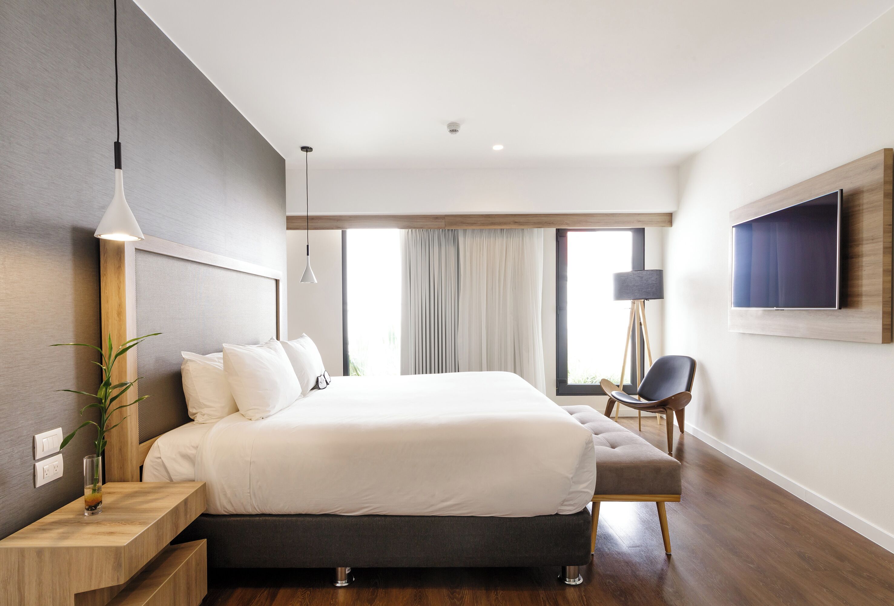 A modern hotel room with a large bed, a bedside table with a lamp, a chair, and a TV mounted on the wall.