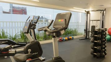 Un gimnasio con equipo de ejercicio, incluyendo cintas de correr y mancuernas, y vista panorámica de la ciudad a través de amplios ventanales.