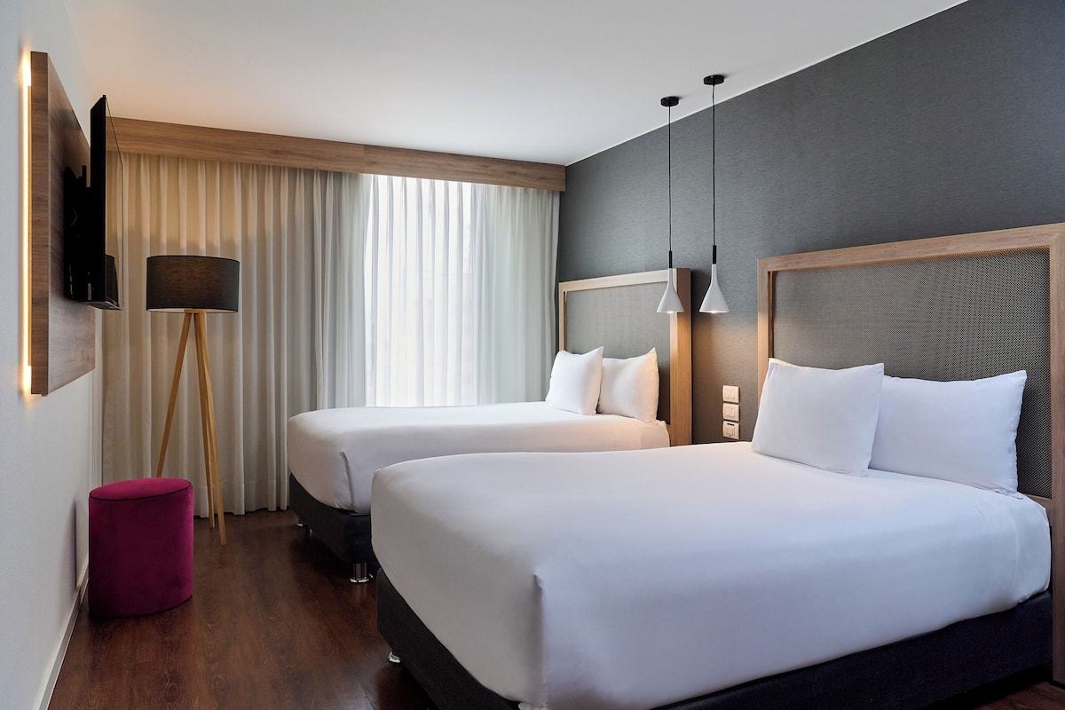 Quarto de hotel com duas camas, televisão na parede, abajur e uma janela grande com cortinas.