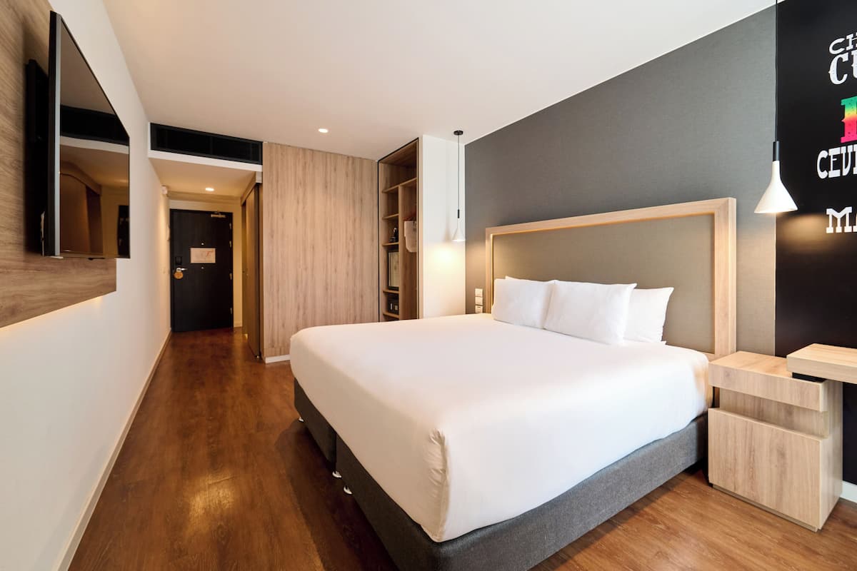 Quarto de hotel moderno com cama grande, piso de madeira, guarda-roupa embutido e mesa de cabeceira.