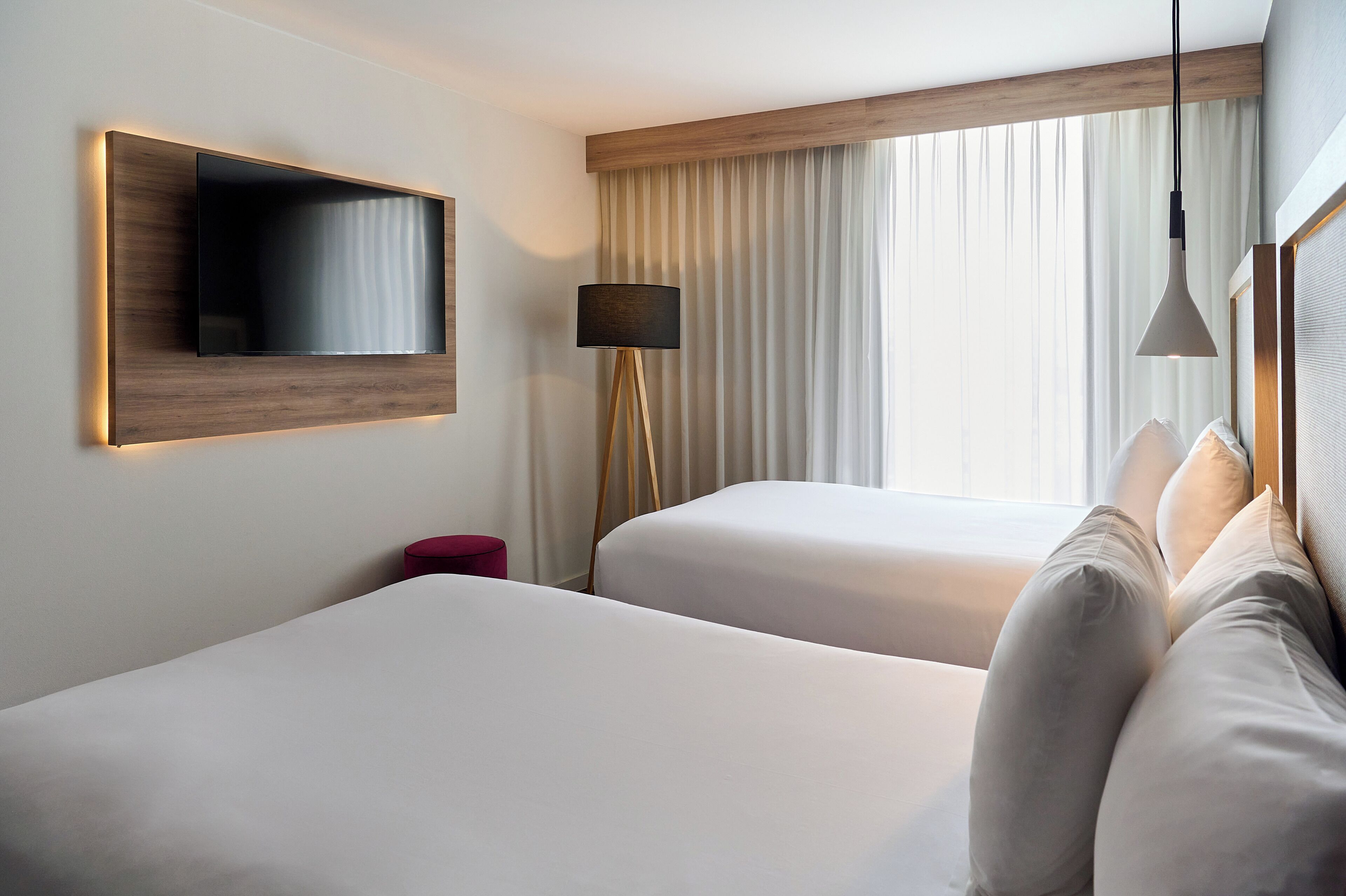 A hotel room with two beds, a flat-screen TV mounted on the wall, a floor lamp, and a window with curtains.
