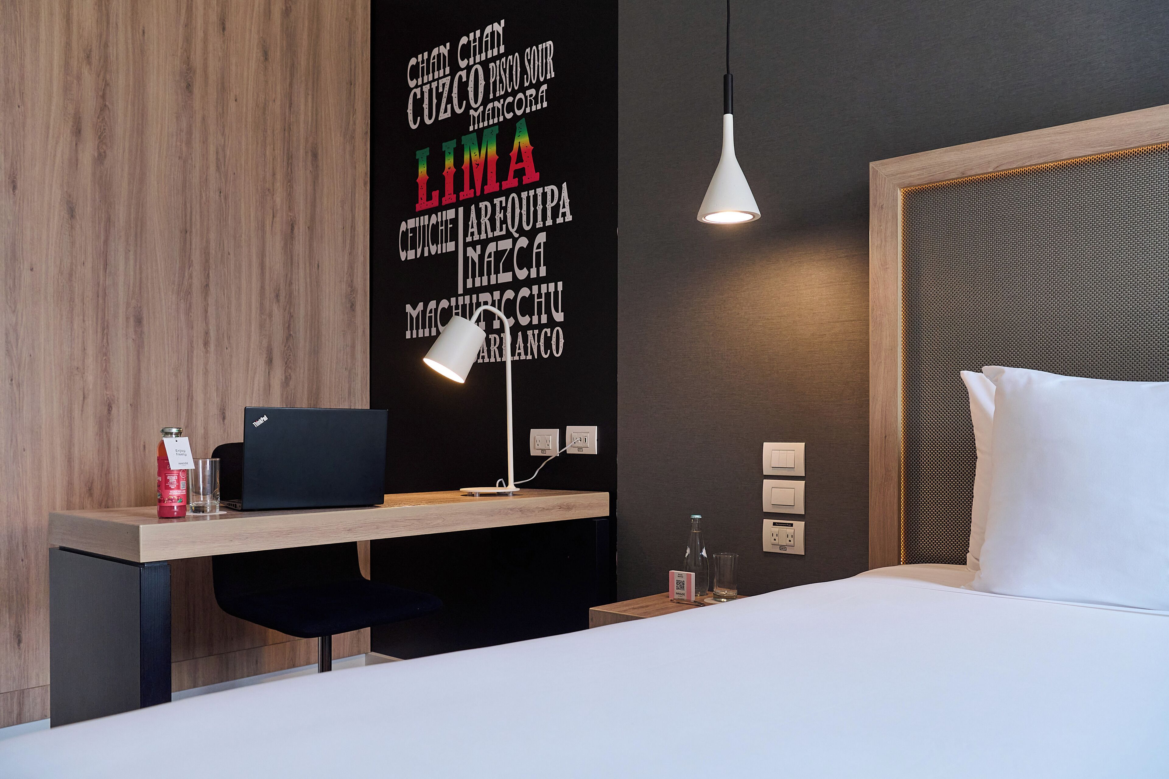 Ein modernes Hotelzimmer mit einem Holztisch, einem Laptop, einer Lampe und einem Bett mit weißen Leinen.