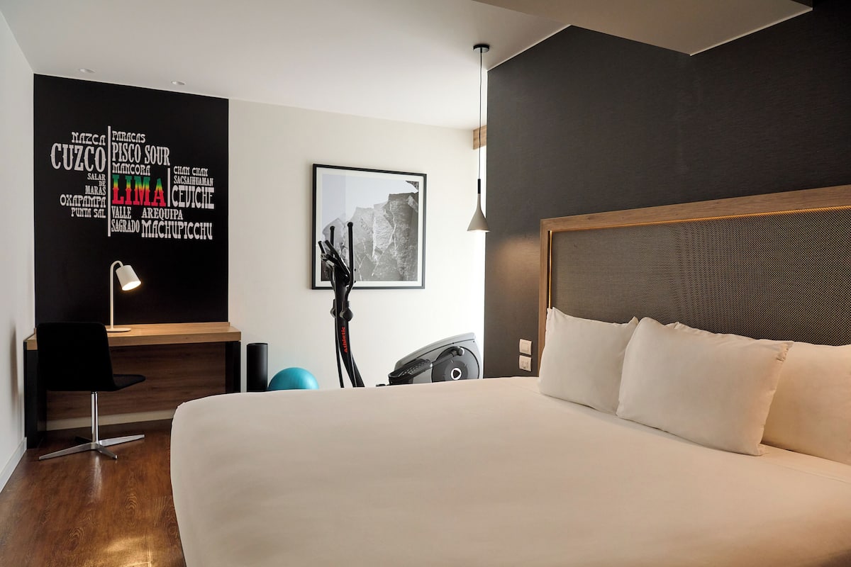 Quarto de hotel moderno com uma cama grande, uma escrivaninha com cadeira, uma bicicleta ergométrica e uma parede com nuvem de palavras.