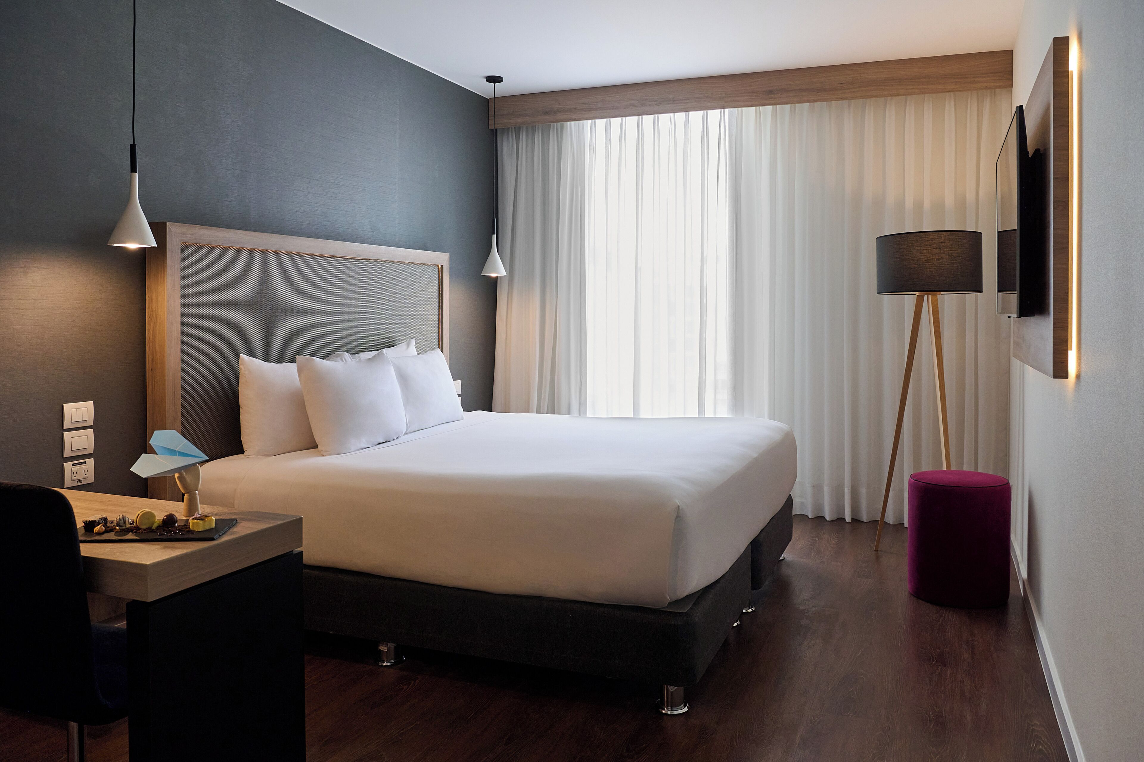 Une chambre d’hôtel moderne avec un grand lit, un bureau avec une lampe, un lampadaire et un téléviseur fixé au mur.