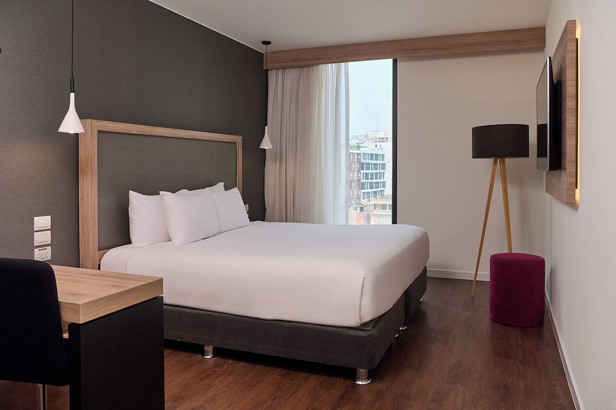 Quarto de hotel moderno com uma cama grande, uma escrivaninha, uma cadeira, um abajur e vista para a cidade pela janela.