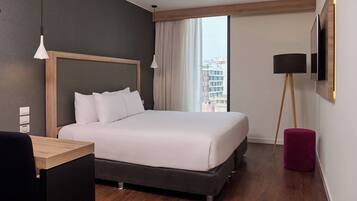 Una habitación de hotel moderna con una cama grande, un escritorio, una silla, una lámpara de pie y vistas de la ciudad a través del ventanal.