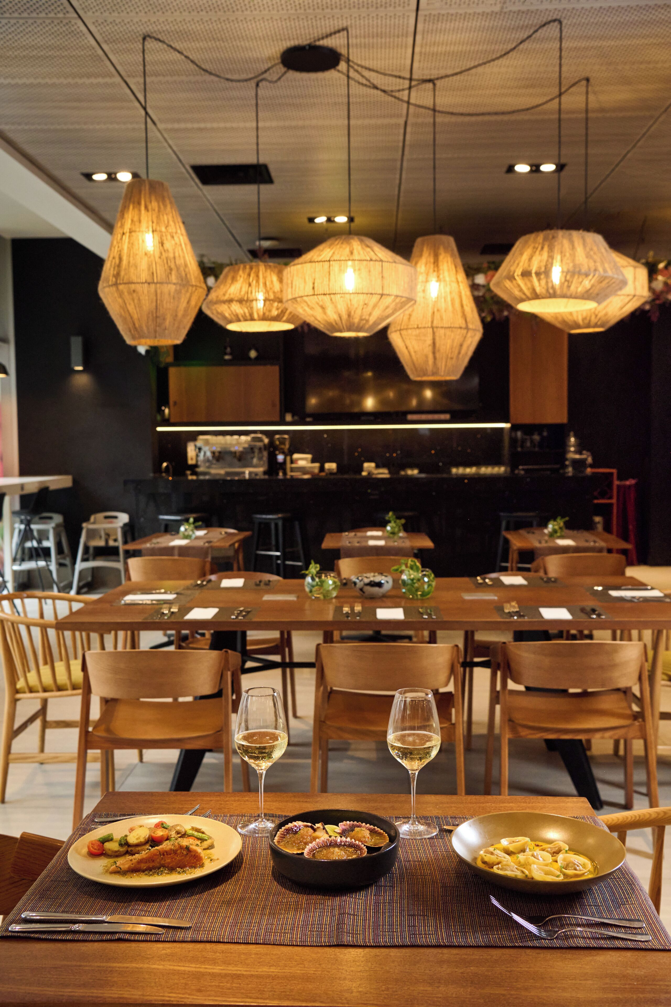 A restaurant with wooden tables and chairs, hanging pendant lights, and a bar area in the background.