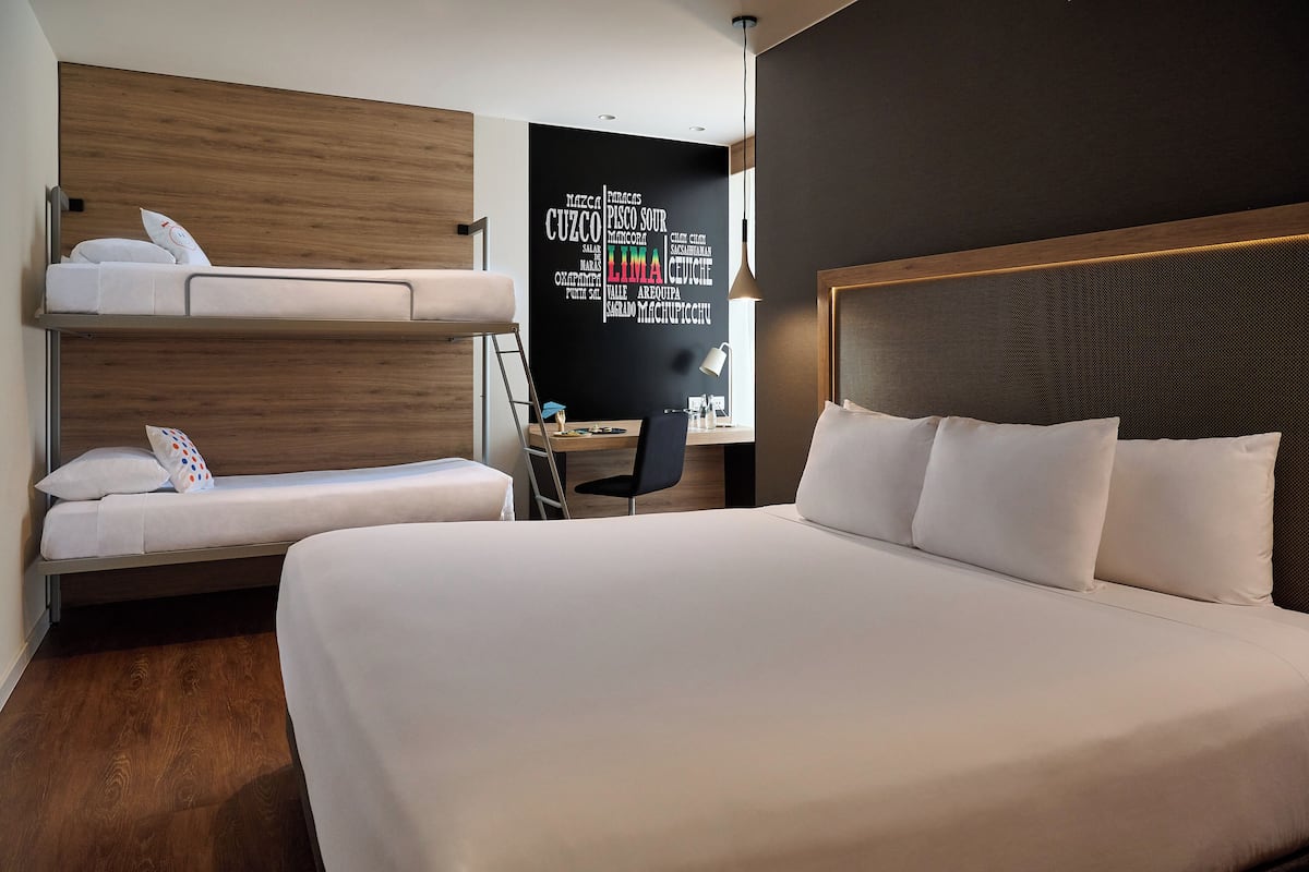 Quarto de hotel com beliche, escrivaninha e cadeira, e uma cama grande com almofadas.