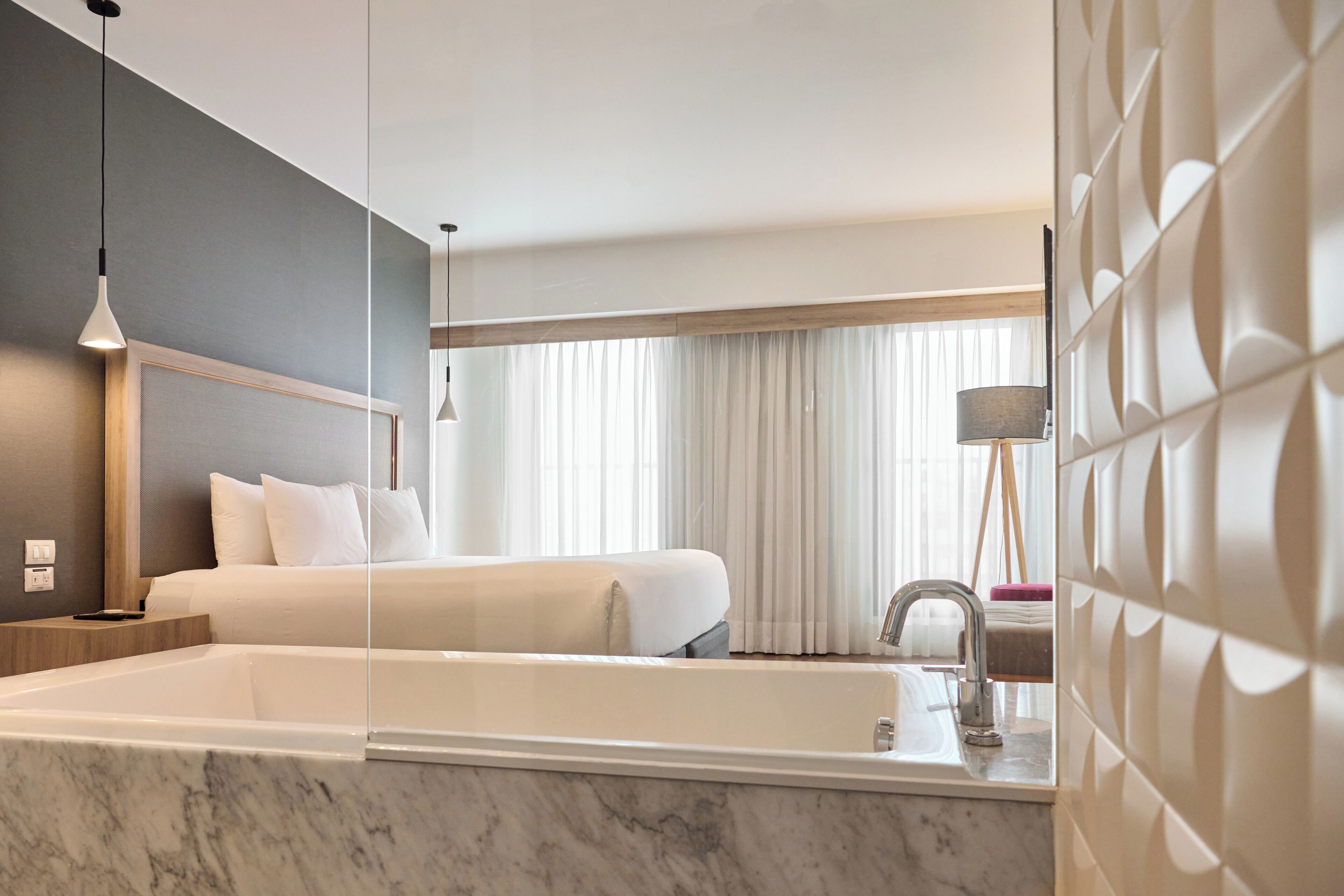 Une chambre d’hôtel moderne dotée d’un grand lit, d’une salle de bain séparée avec baignoire et d’une grande fenêtre avec rideaux.