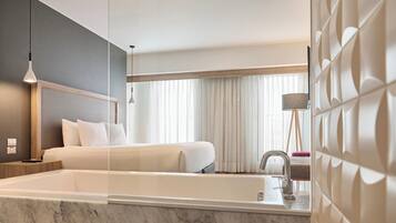 Une chambre d’hôtel moderne dotée d’un grand lit, d’une salle de bain séparée avec baignoire et d’une grande fenêtre avec rideaux.