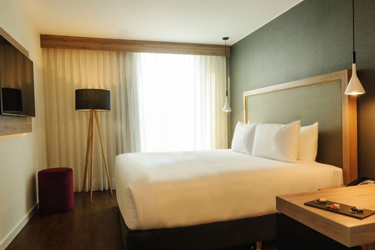 Quarto de hotel com uma cama grande, uma mesinha de cabeceira com um controle remoto, uma luminária e vista para o exterior.