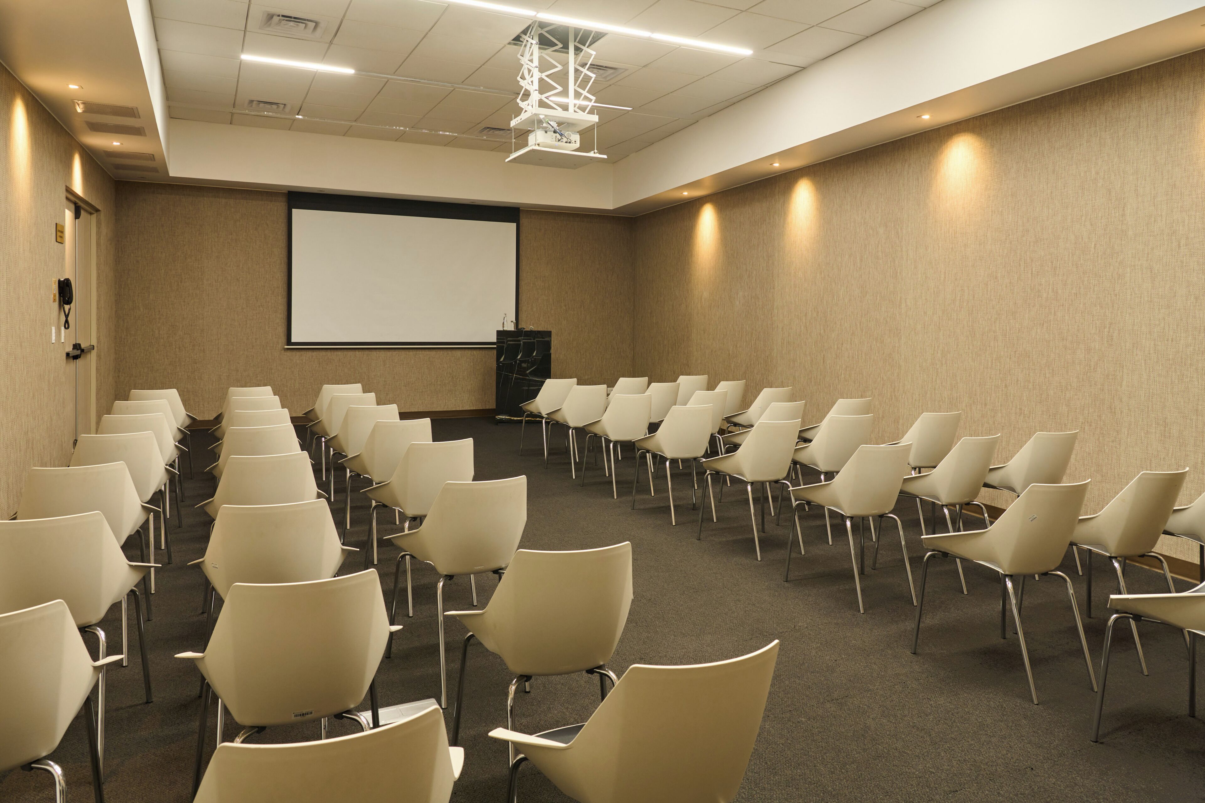 Un salón de conferencias con filas de sillas mirando hacia una pantalla de proyección, un atril y piso alfombrado.