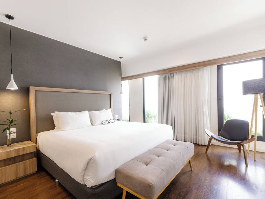A modern hotel room with a large bed, a bedside table, a bench, a chair, and a lamp.