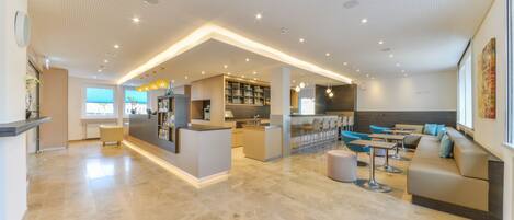 A modern hotel lobby with a central reception desk, seating area, and a bar.