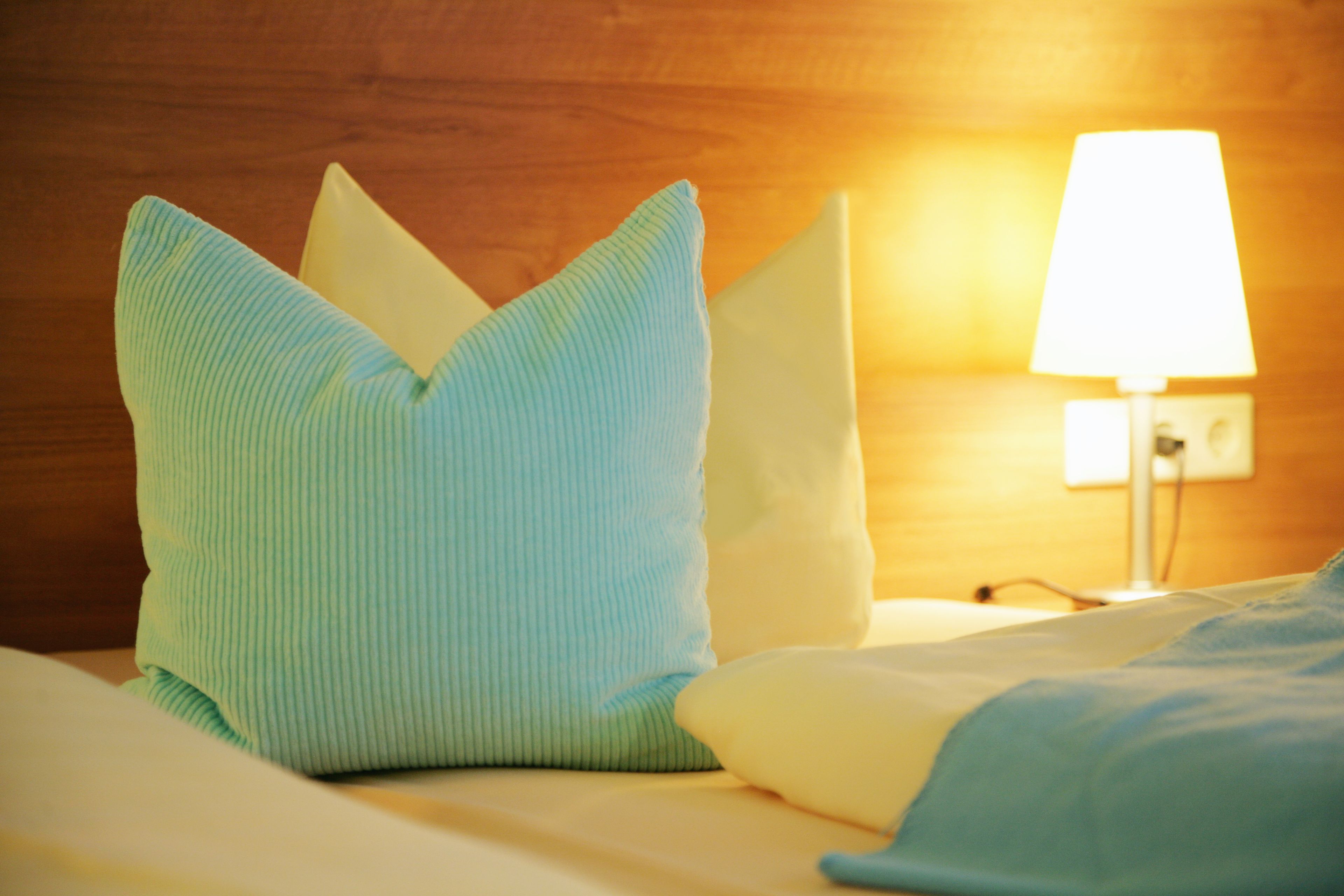 A bed with a blue pillow and a white pillow, a lamp on the wall, and a wooden headboard.