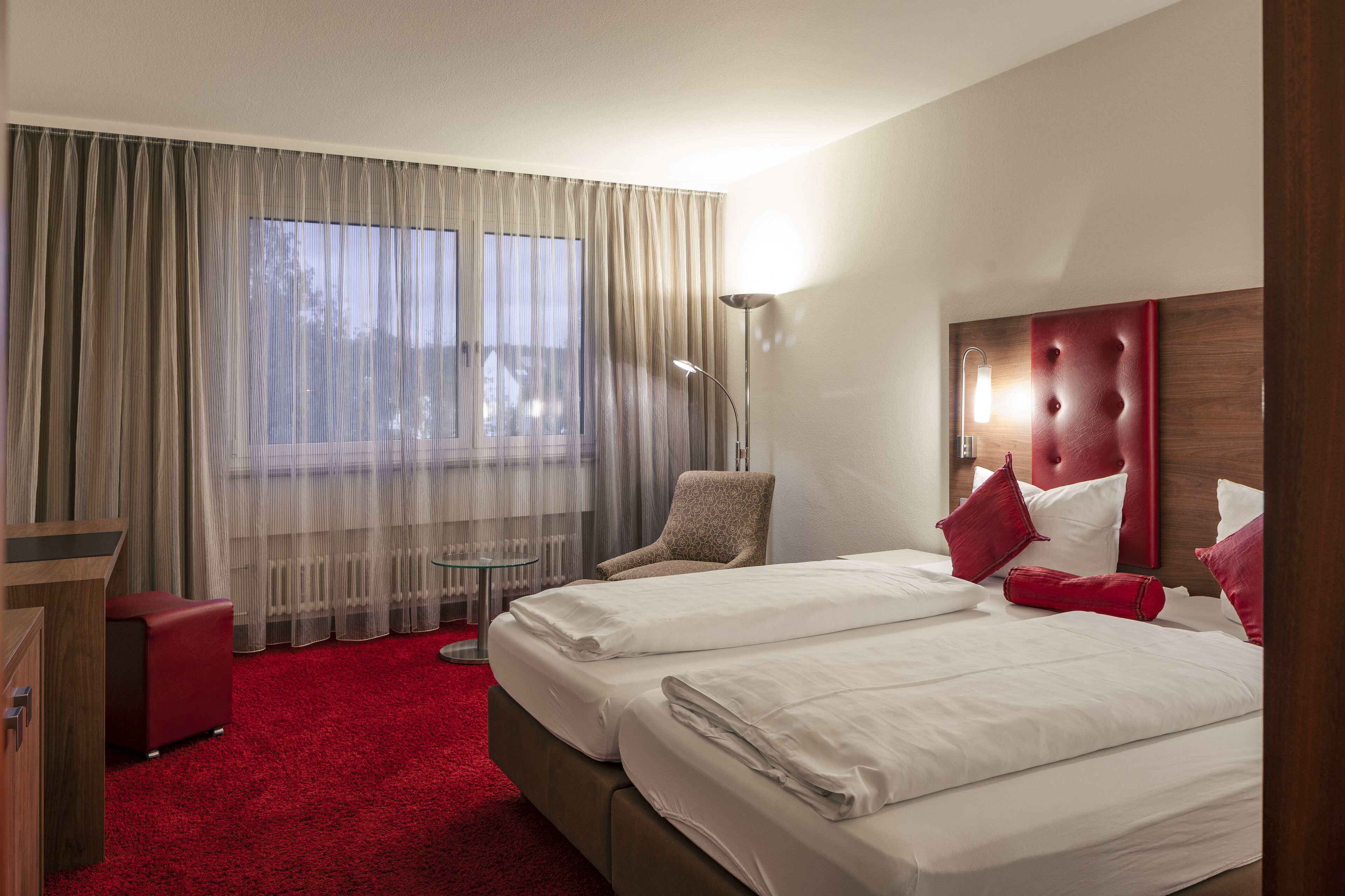 A hotel room with two beds, a red armchair, a desk, and a large window with sheer curtains.