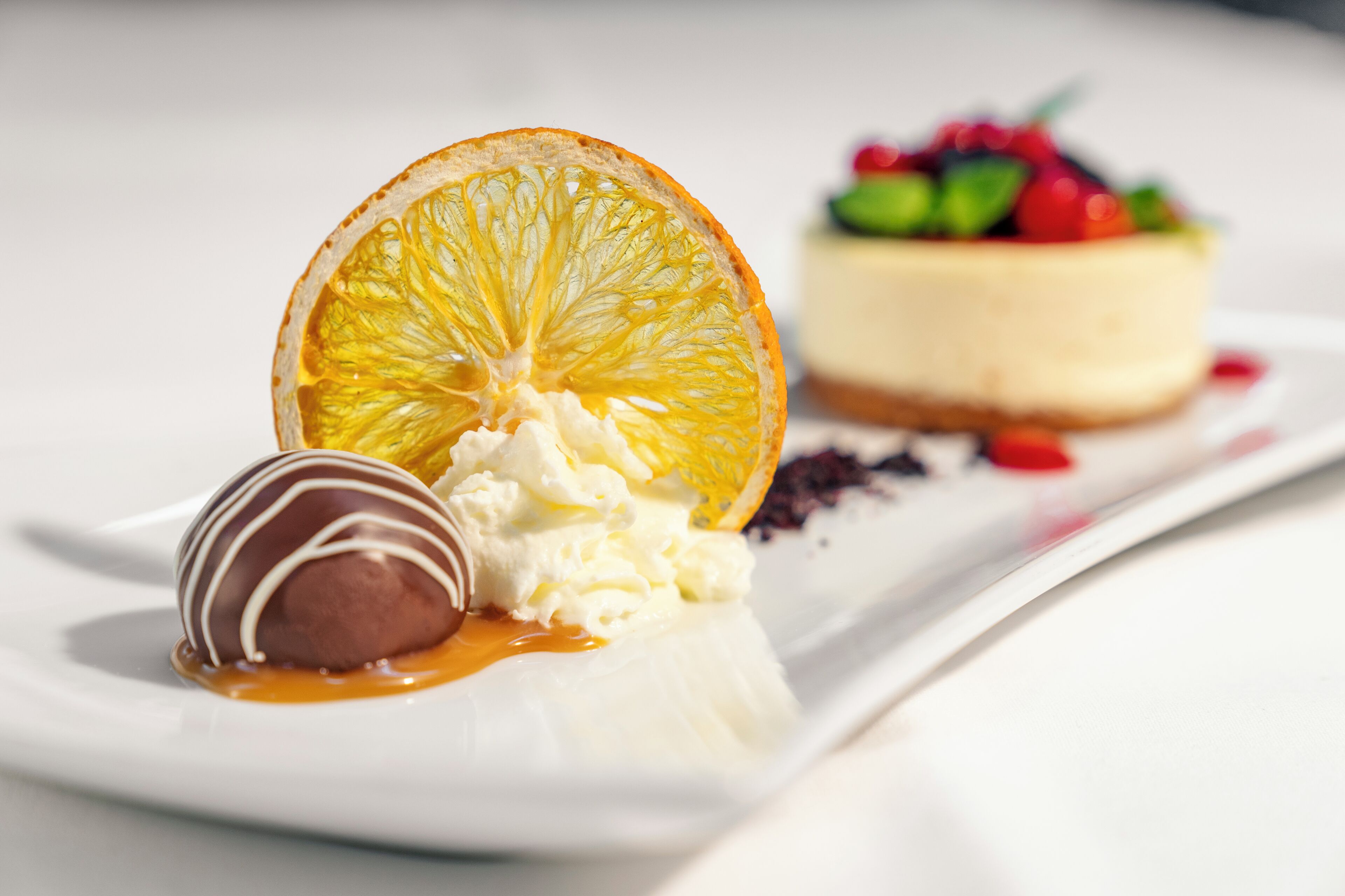 Plato de postre con una esfera de chocolate, crema batida y salsa de caramelo, acompañado de una rodaja de naranja y un trozo de pastel de queso con frutos rojos.
