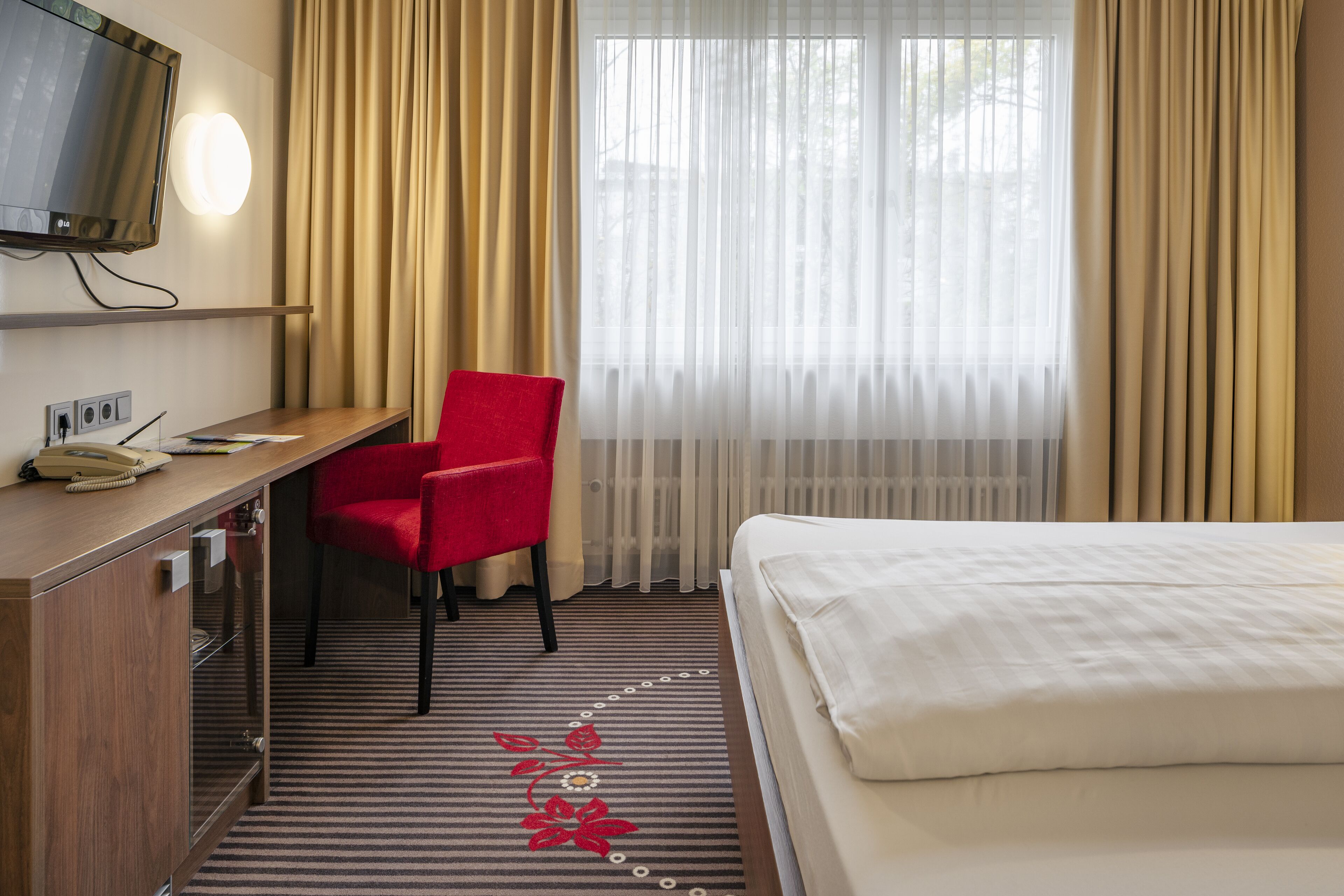 Una habitación de hotel con una cama, un escritorio con teléfono, una silla roja, un televisor y una ventana con cortinas.