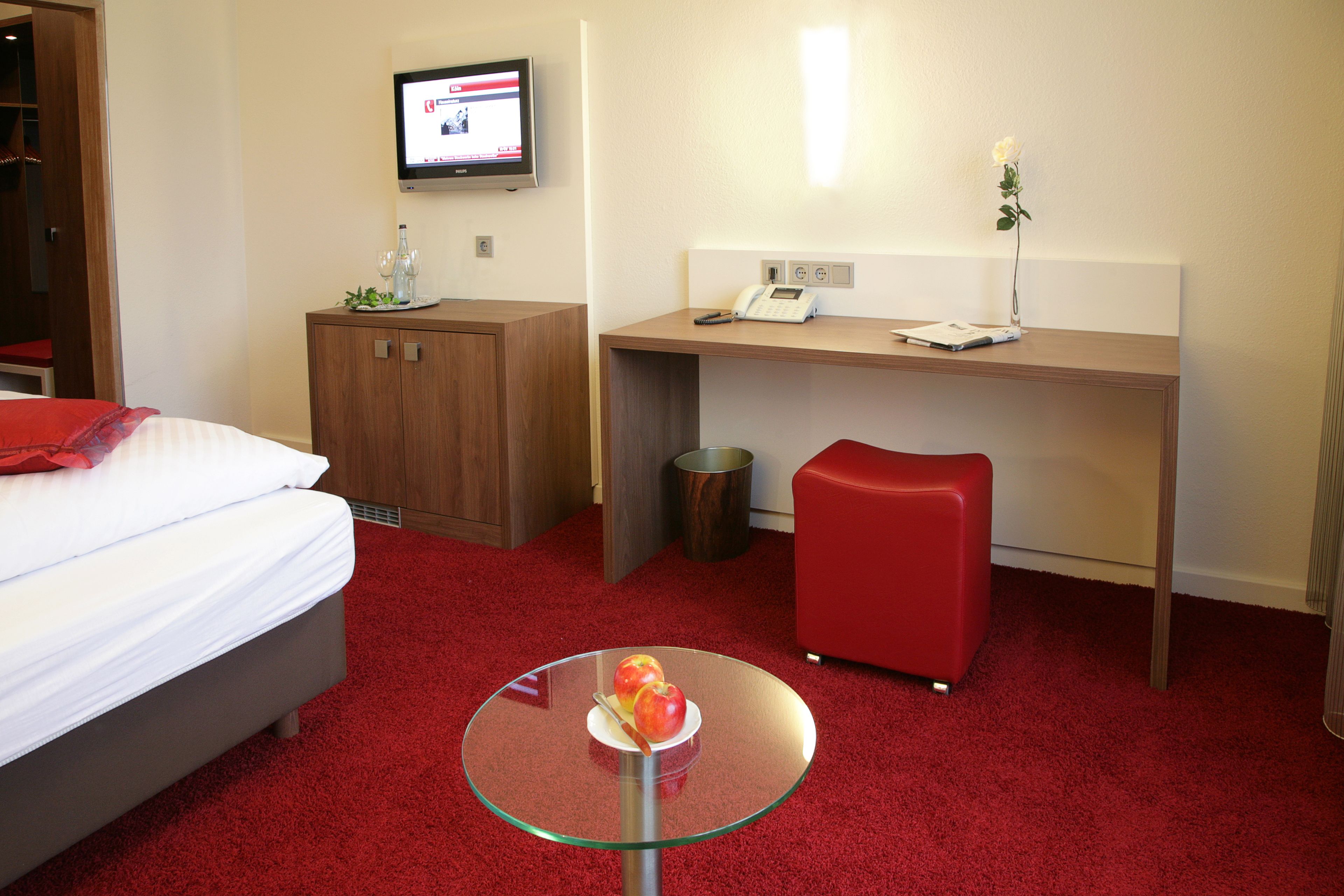 Una habitación de hotel con una cama, un escritorio, una pequeña mesa con manzanas, una silla roja, un televisor en la pared y un cubo de basura.