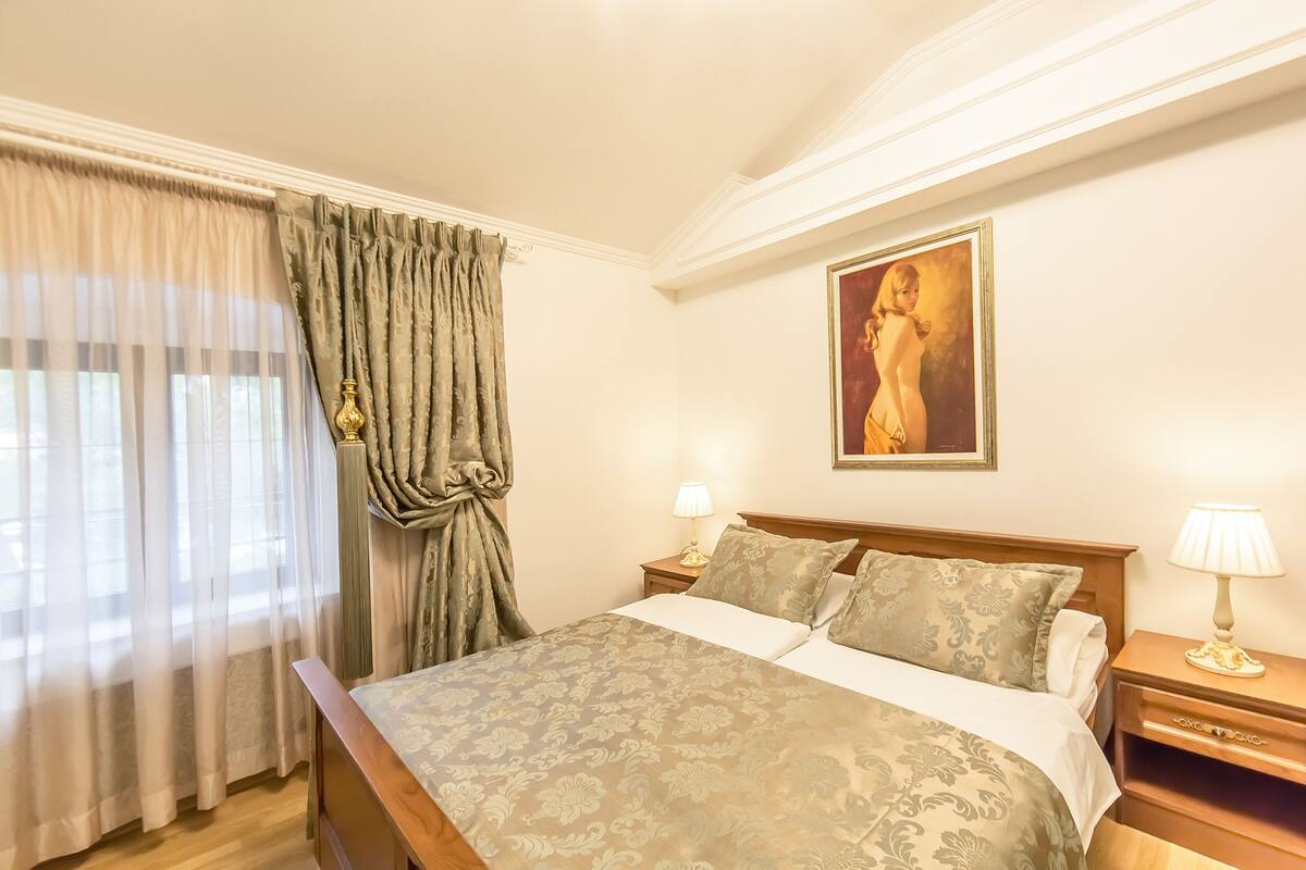 A hotel room with a bed, curtains, a painting, and bedside lamps.