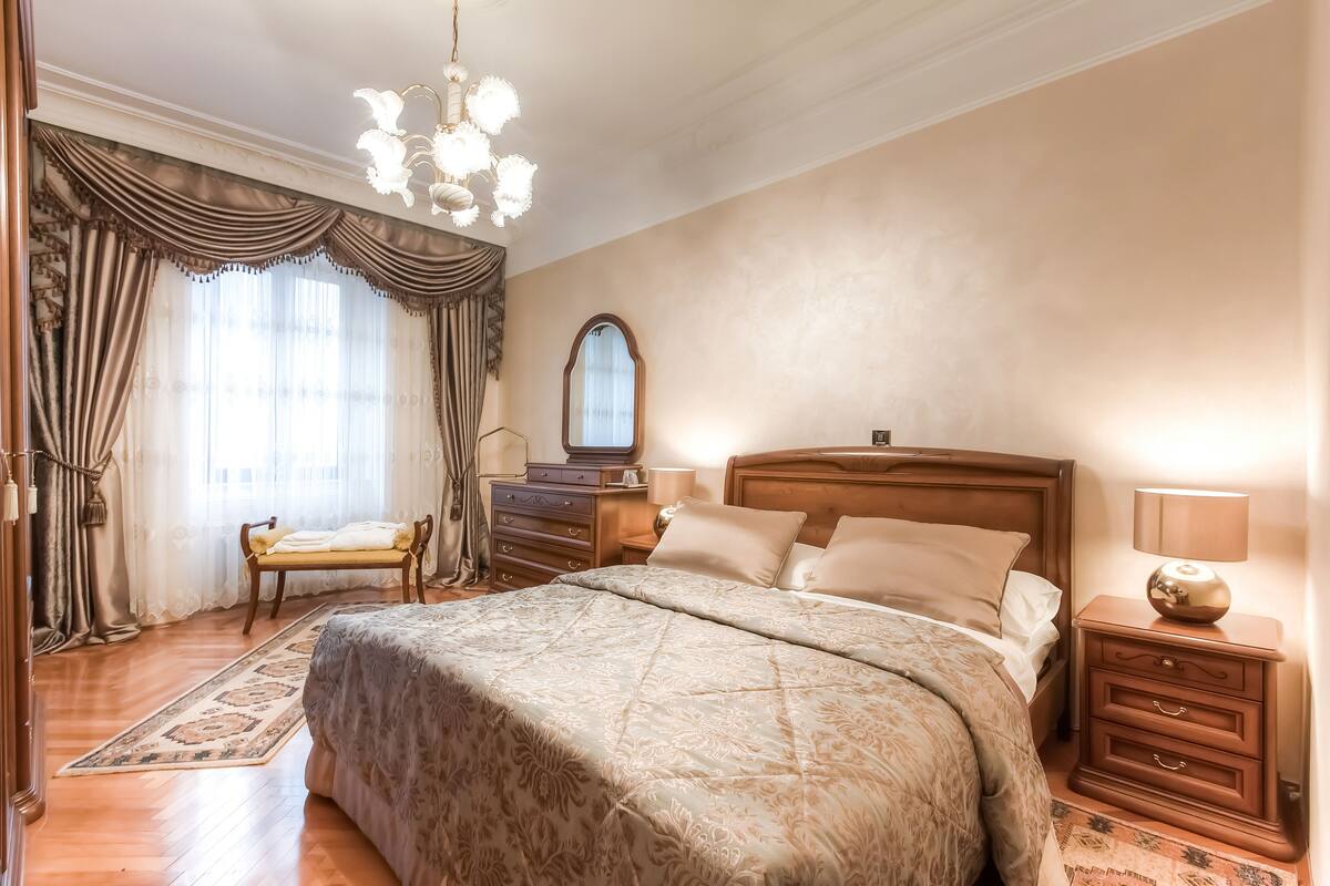 A bedroom with a large bed, wooden nightstands, a bench, a chandelier, and a large mirror.