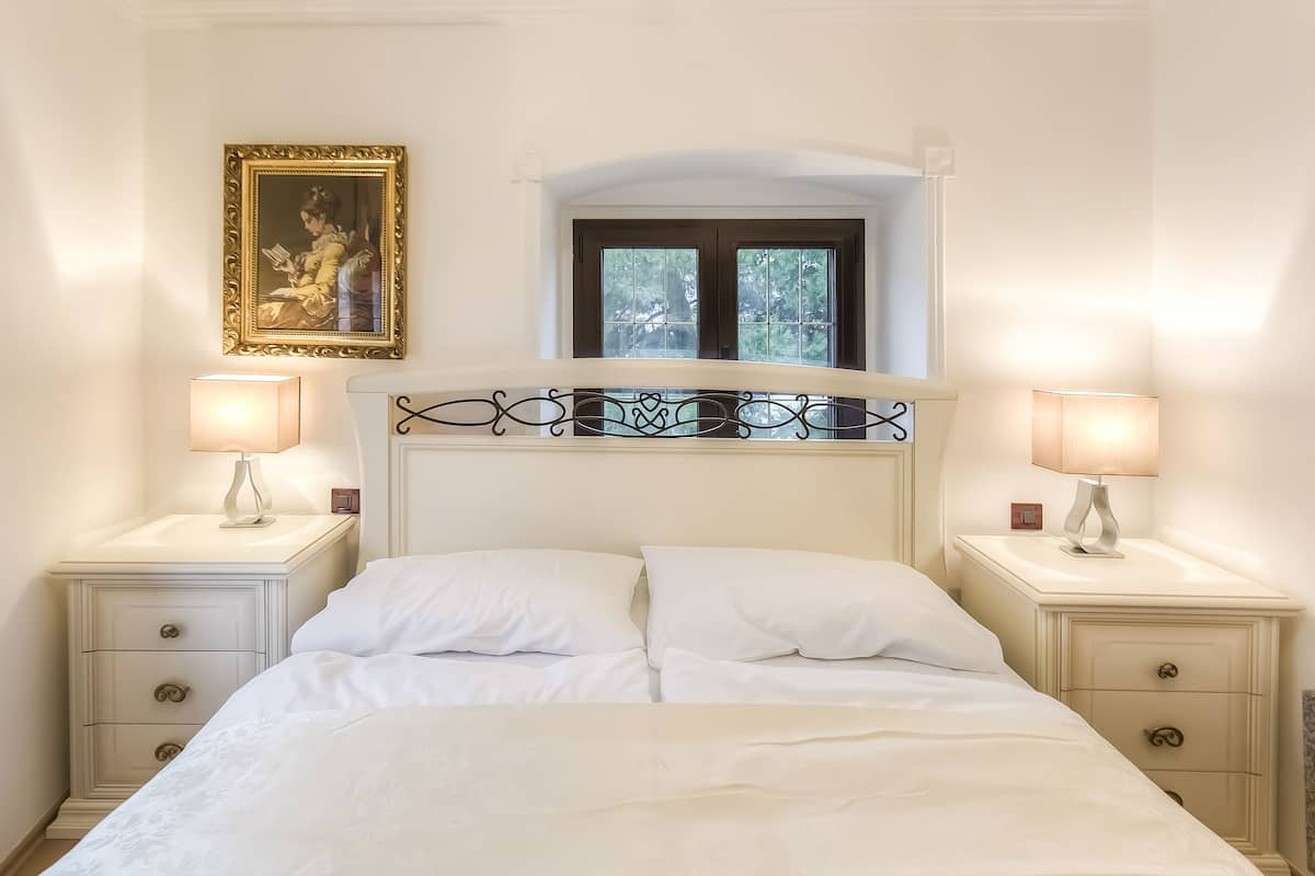 A bedroom with a bed, two bedside tables, a nightstand, a window, and a framed picture on the wall.