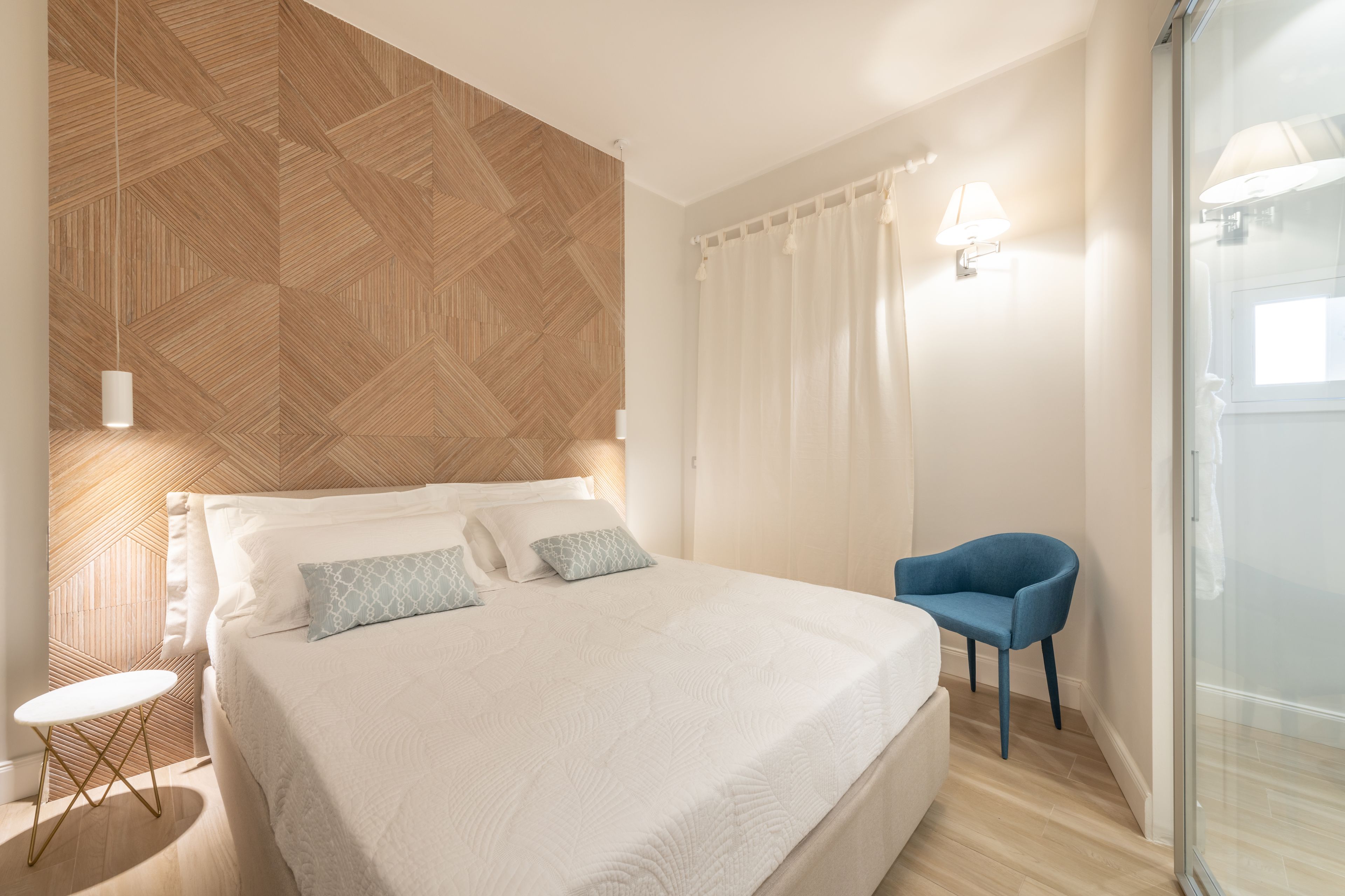 A bedroom with a bed, a blue chair, a small table, and a glass shower area.