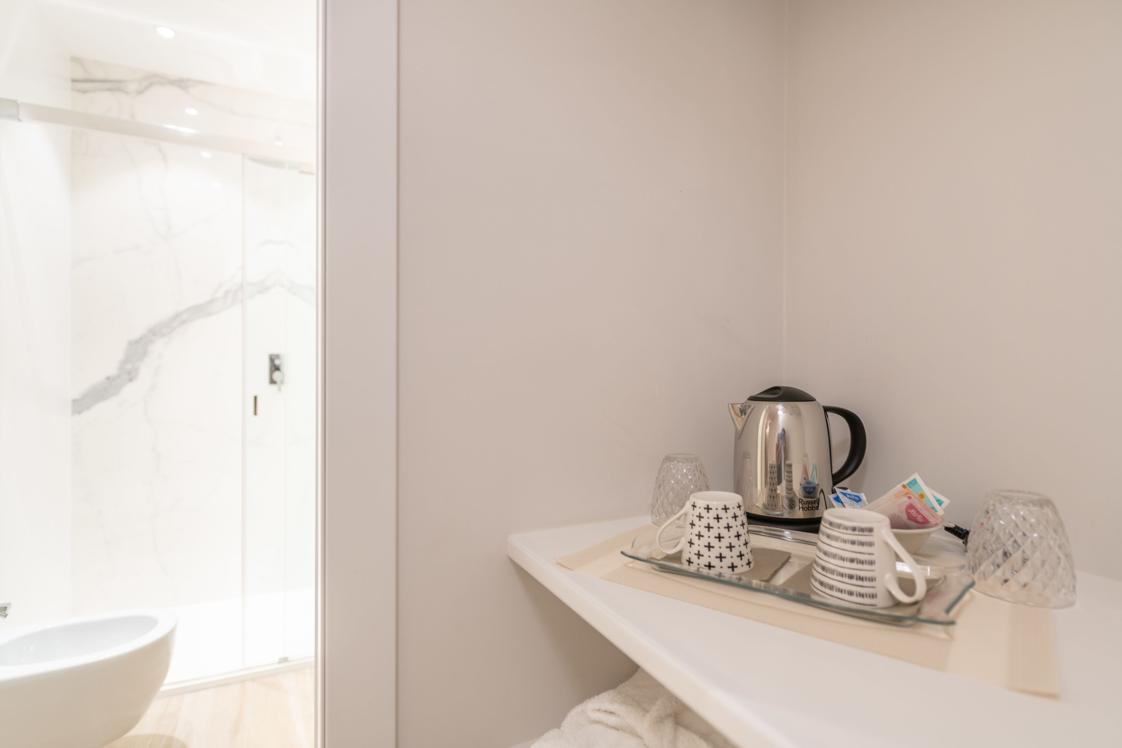 Une salle de bain avec un lavabo blanc, une douche et un plan de travail blanc sur lequel se trouvent une bouilloire, des tasses et des verres.
