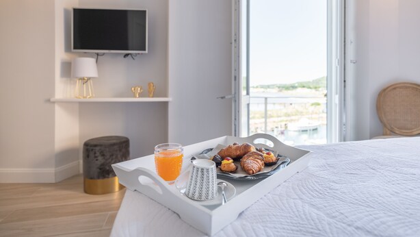 Una bandeja de desayuno bien dispuesta con croissants y zumo de naranja en una cama de habitación de hotel, con televisión y vistas al exterior.