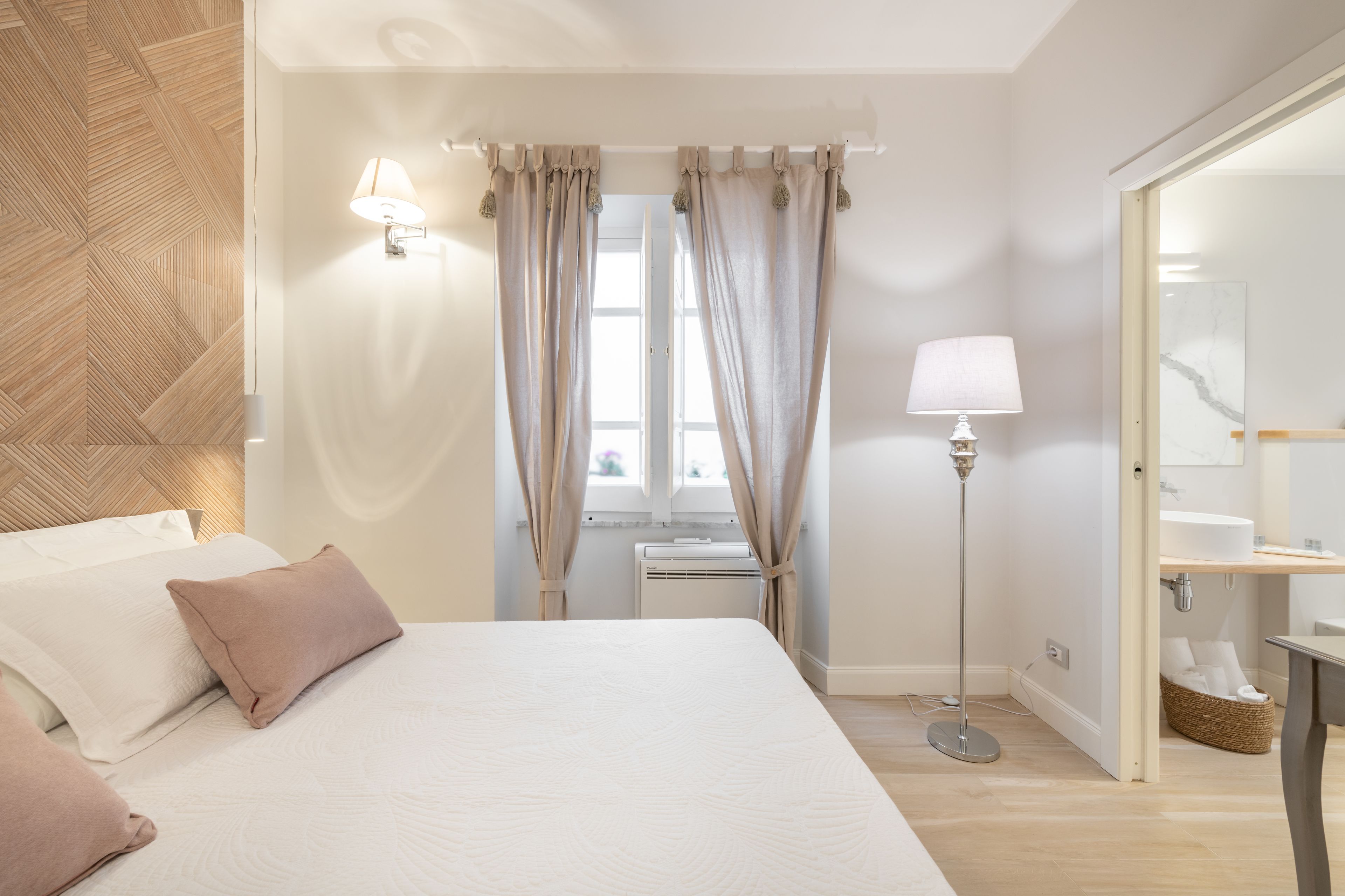 Une chambre à coucher avec un lit, des oreillers, une lampe de chevet, une fenêtre avec des rideaux, un radiateur et un miroir.