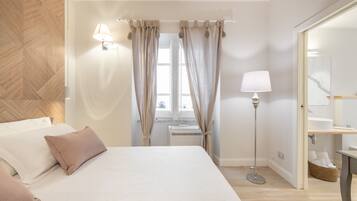 Une chambre à coucher avec un lit, des oreillers, une lampe de chevet, une fenêtre avec des rideaux, un radiateur et un miroir.