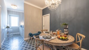 Un comedor con una mesa redonda dispuesta para cuatro personas, rodeada de sillas de madera. Sobre la mesa hay una planta en maceta y un candelabro cuelga del techo. El piso presenta un patrón geométrico y una pared de acento gris.