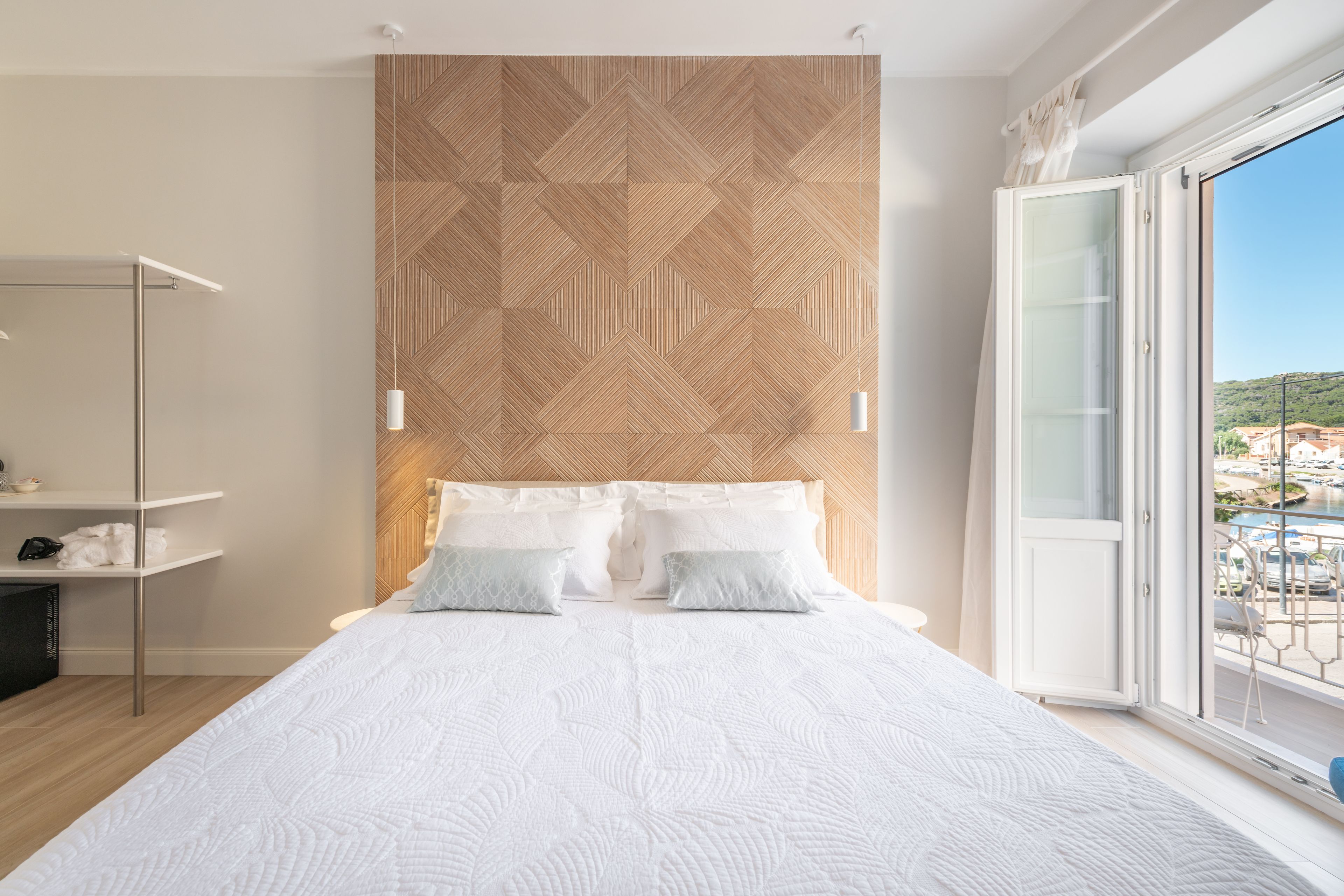 A modern bedroom with a large bed, a geometric patterned headboard, a bedside table with a lamp, and a view of a marina through a sliding glass door.