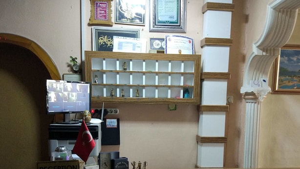 Un comptoir d’accueil avec une enseigne indiquant « RÉCEPTION » et « OTEL PETEK ». Des certificats et des récompenses encadrés sont accrochés au mur derrière le comptoir.