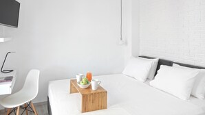 Ett modernt hotellrum med en vit tegelvägg, en säng med vita sängkläder, ett träbord vid sängen med en skål med frukt och ett glas apelsinjuice, en vit stol och ett skrivbord med en bärbar dator och en bok.