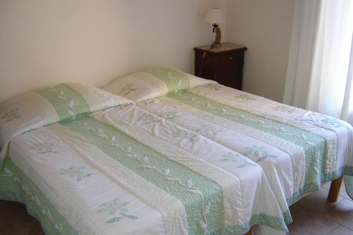 A bed with a green and white floral bedspread, a bedside table with a lamp, and a window with curtains.