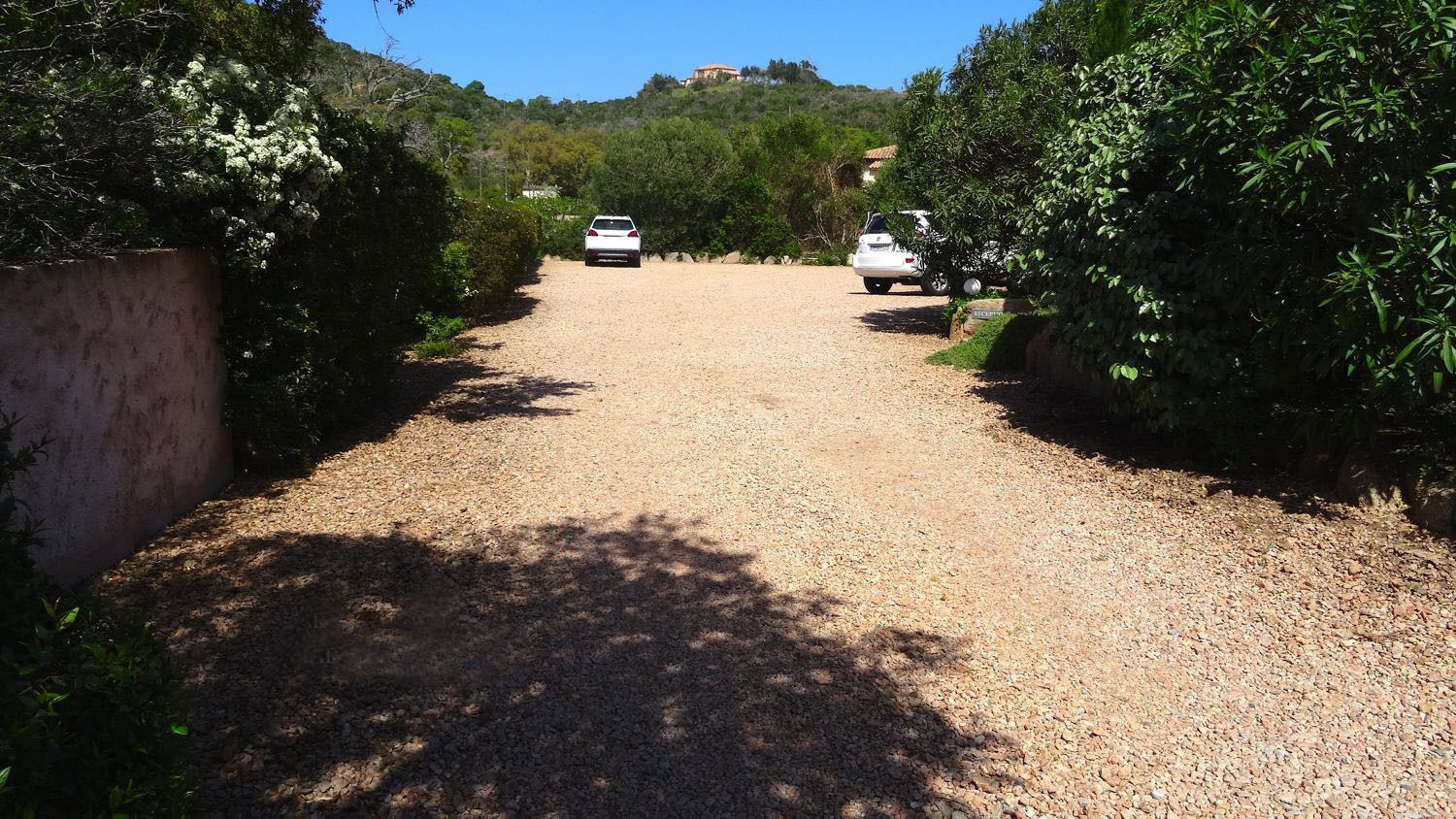 Une allée de gravier mène à une voiture garée, entourée de verdure et avec une colline en arrière-plan.