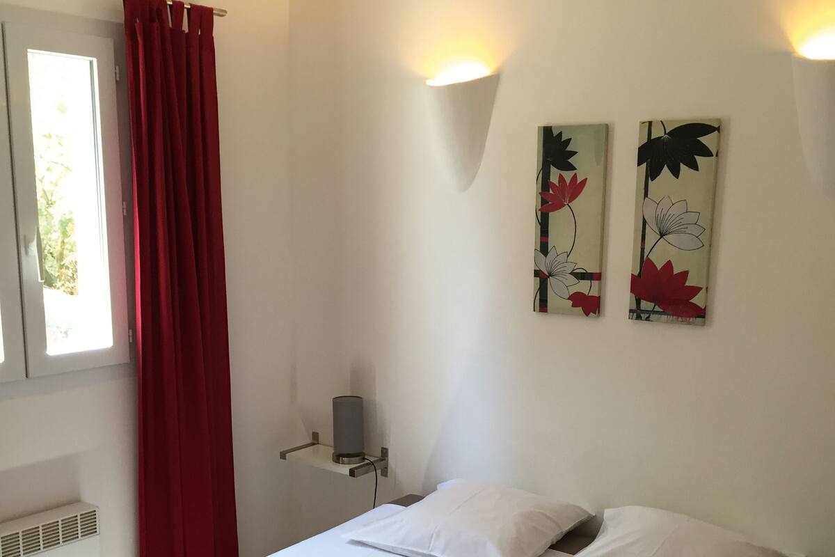 A neatly made bed with white linens, a red-striped blanket, and two pillows. There are two wall-mounted light fixtures and two floral wall art pieces. A window with a red curtain is visible, and a small heater is mounted on the wall.