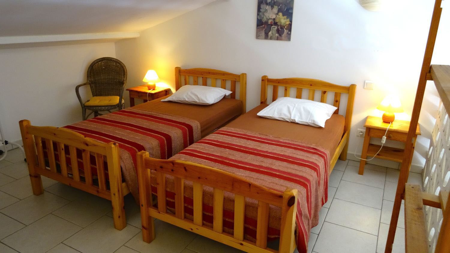A room with two wooden beds, each with a red and white striped blanket, and a single chair with a yellow cushion. There are two bedside tables with lamps.