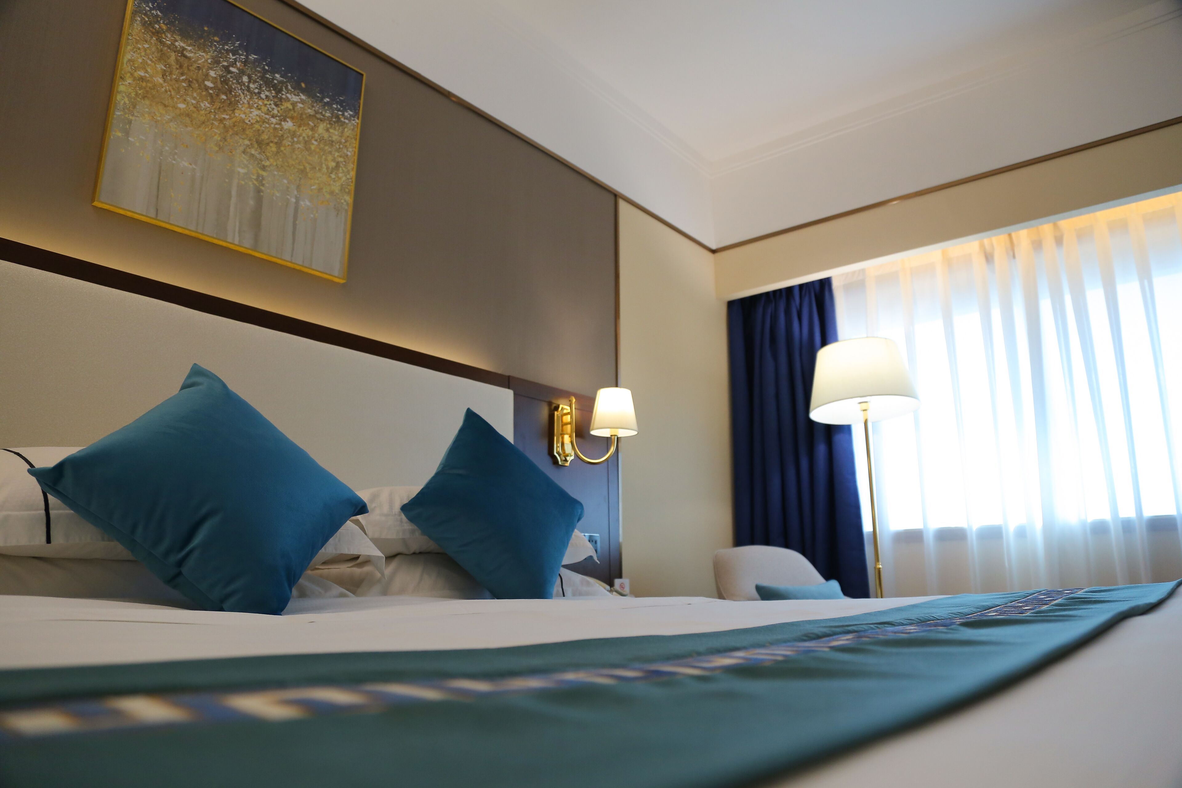 Una habitación de hotel con una cama, dos almohadas azules, una lámpara de pared y un cuadro.