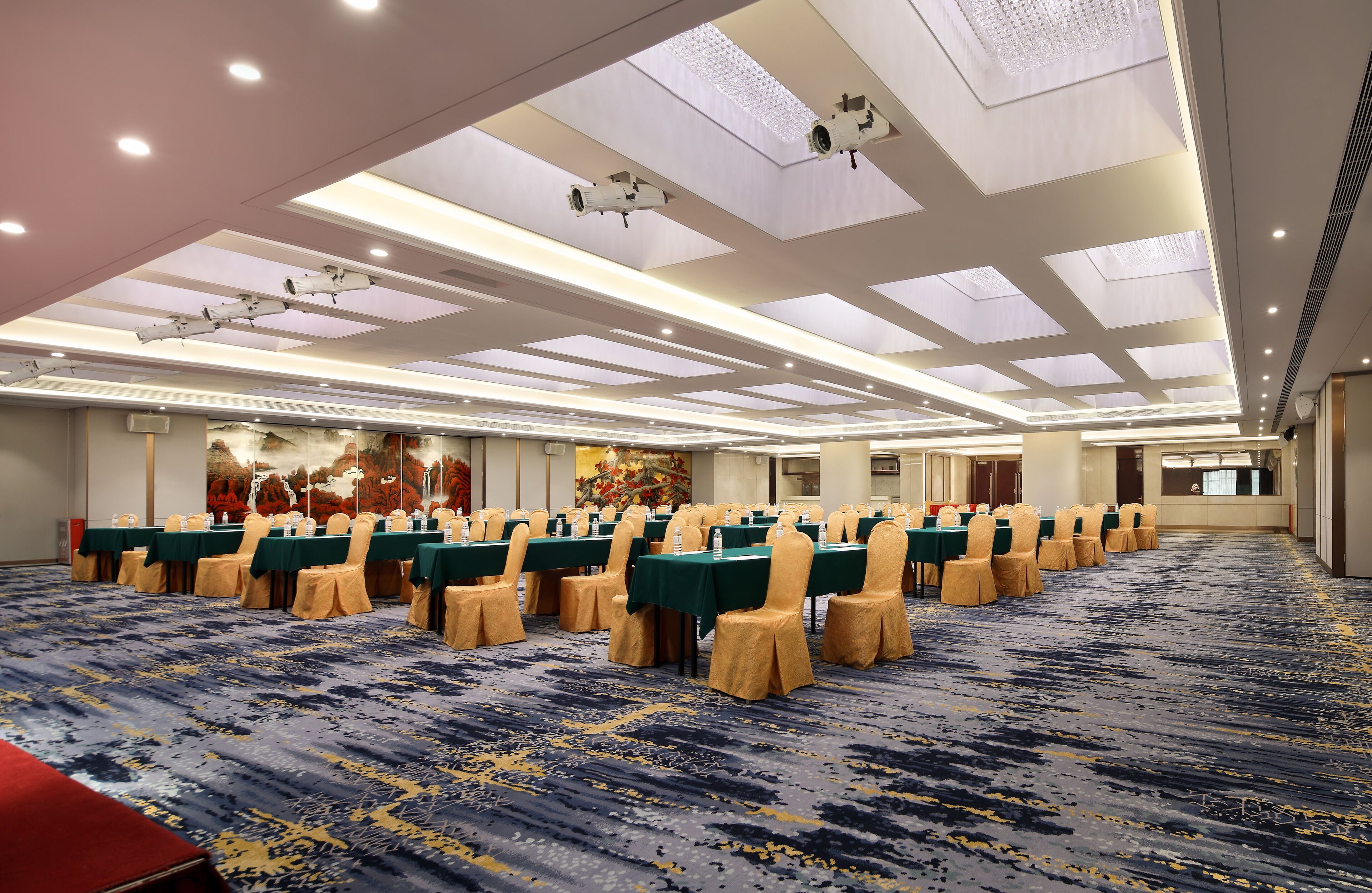 Un salón de conferencias con filas de mesas y sillas, una alfombra estampada y un techo con iluminación empotrada y tragaluces.