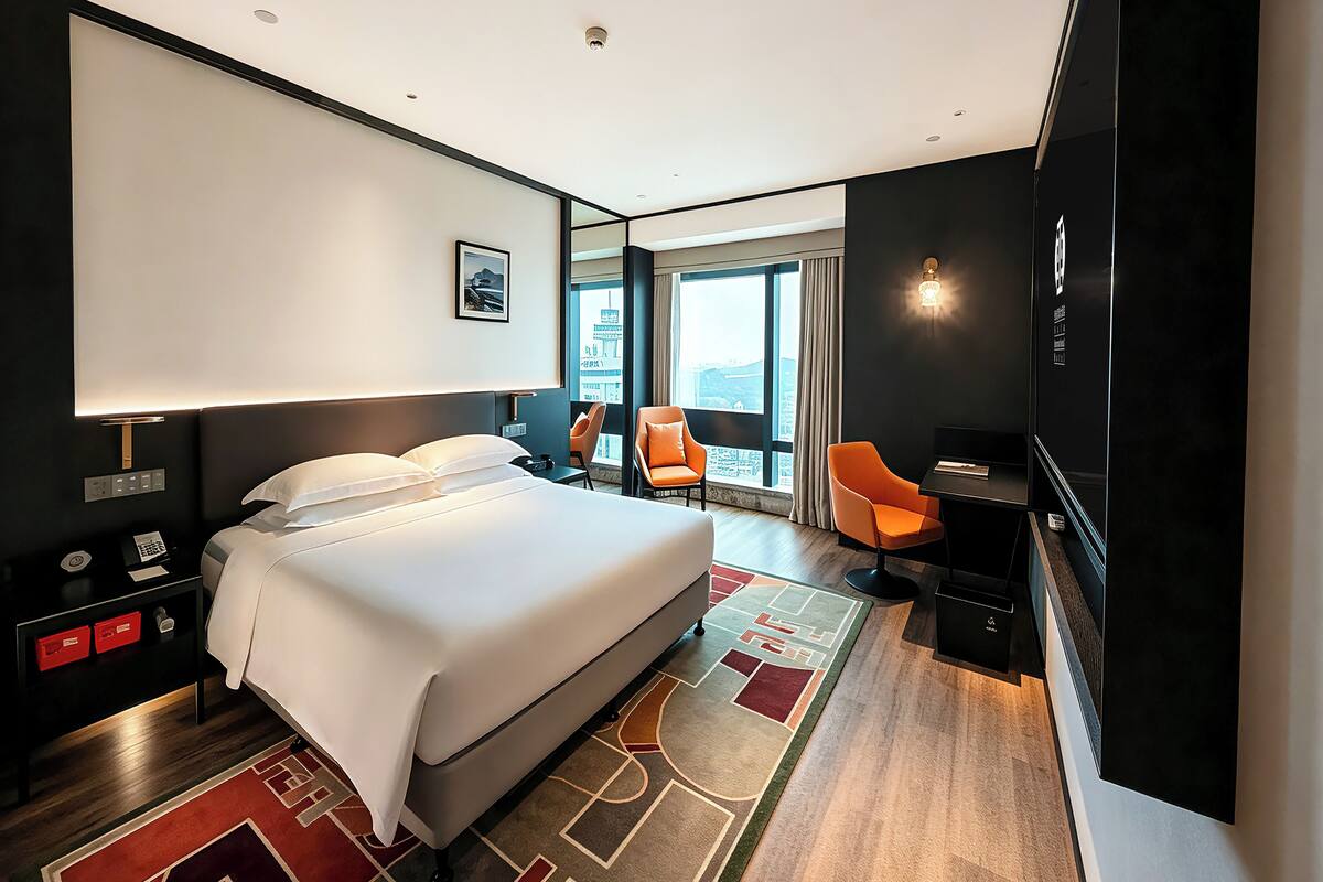 Een moderne hotelkamer met een groot bed, een bureau, twee stoelen, een bureaulamp, een nachtkastje en uitzicht op de stad door grote ramen.
