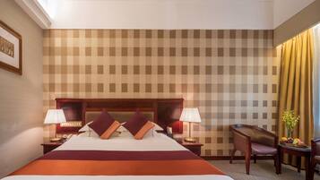 Habitación de hotel con una cama grande, dos mesitas de noche con lámparas, un cabecero de madera, un escritorio con una silla y una ventana con cortinas.