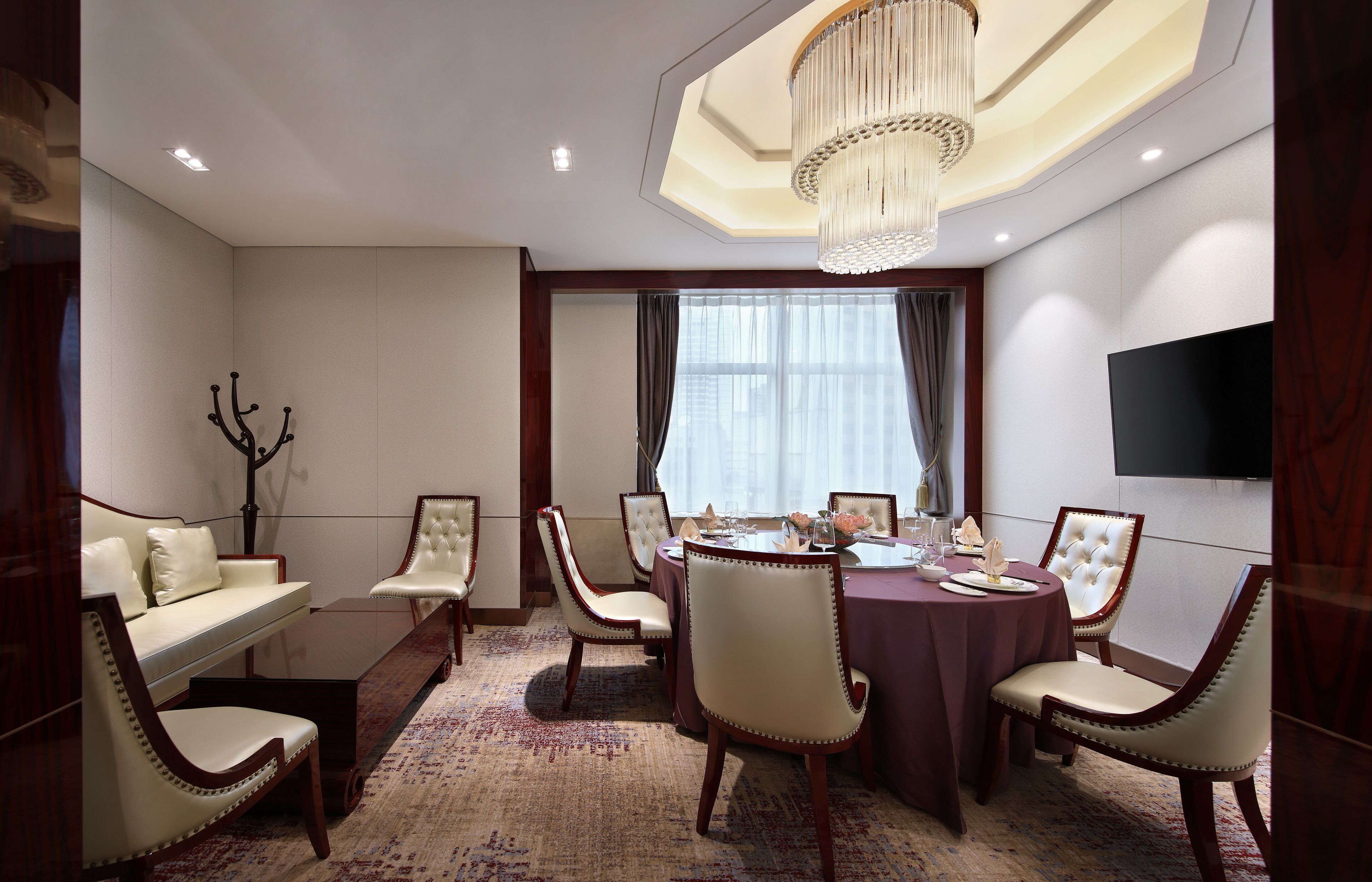 A dining room with a round table, six chairs, a chandelier, a sofa, and a television on the wall.