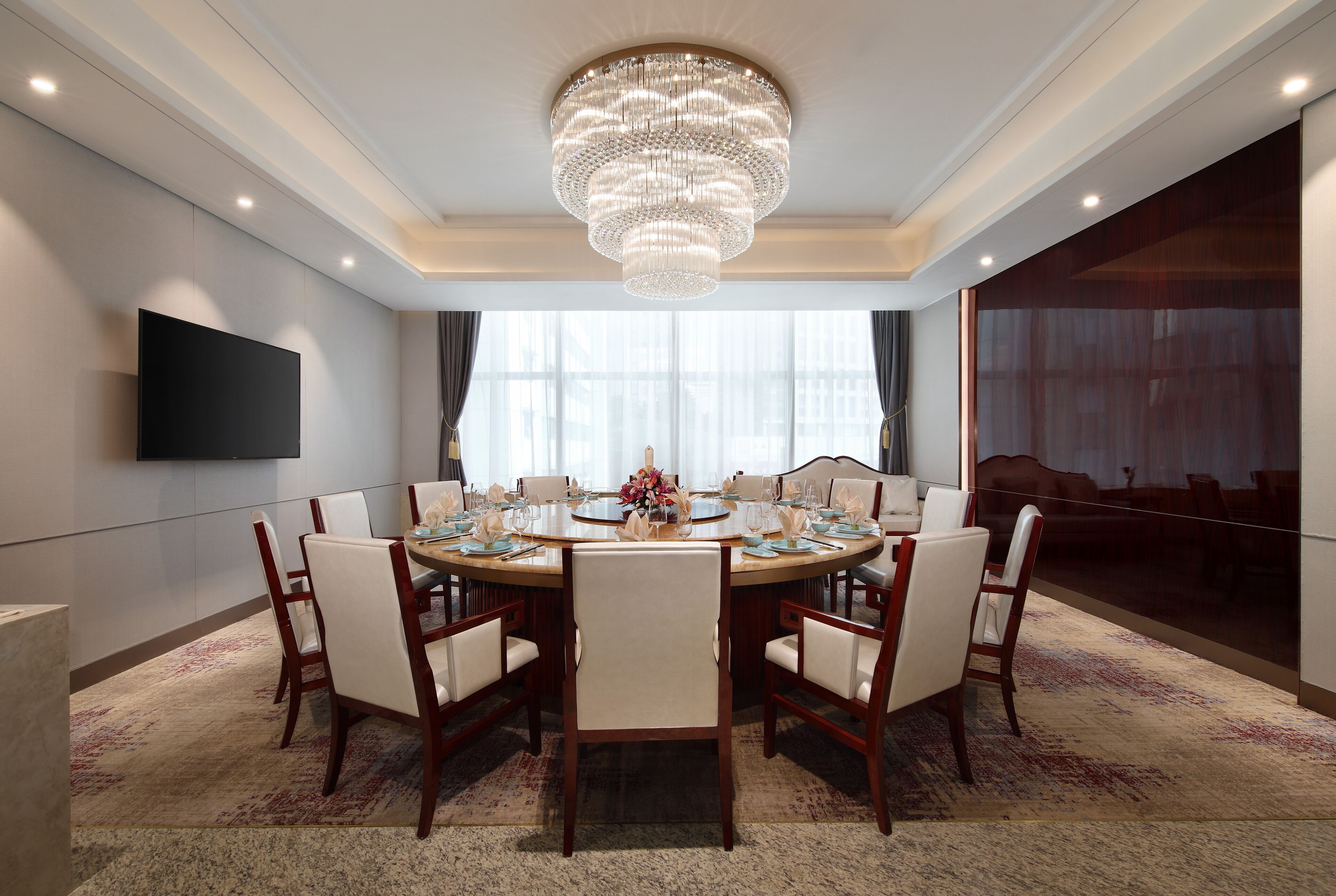A conference room with a round table, chairs, a chandelier, a flat-screen TV, and a large window.